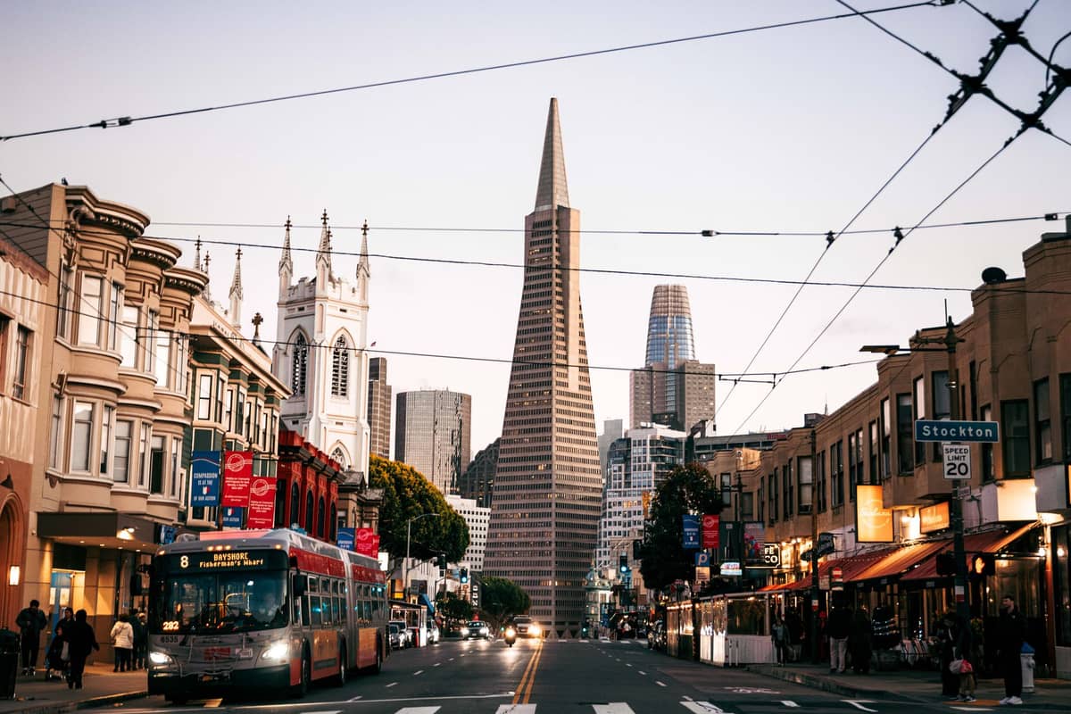 Jalan kota San Francisco dengan bus Muni ikonik di depan, dikelilingi gedung-gedung perkotaan dan cakrawala Transamerica Pyramid yang mudah dikenali.