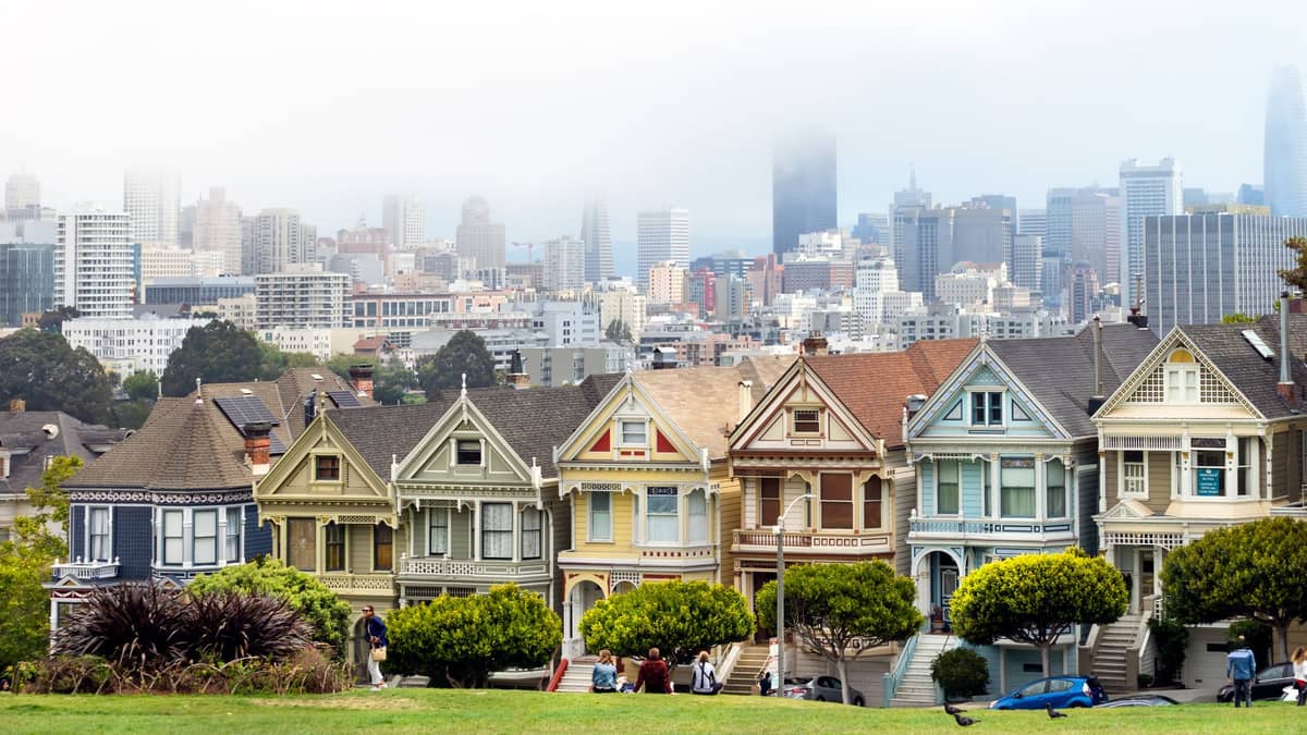 Painted Ladies viktorianische Häuser am Alamo Square mit der Skyline von San Francisco und dem Park im Hintergrund an einem teils bewölkten Tag.