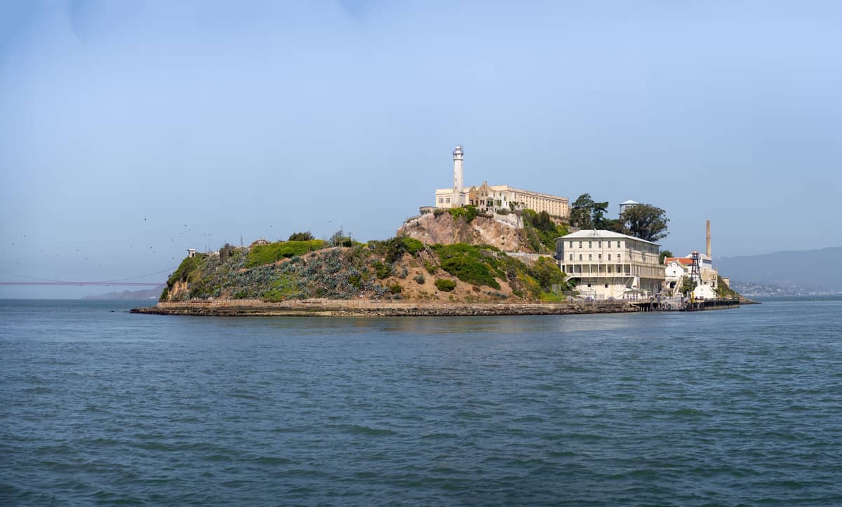 Panoramablick auf Alcatraz Island in der San Francisco Bay, mit den historischen Gefängnisgebäuden und dem Leuchtturm, umgeben von Wasser unter einem klaren blauen Himmel.