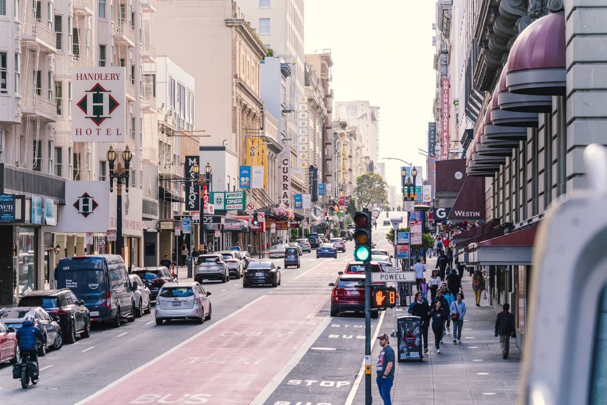 Ruchliwa scena uliczna w centrum San Francisco z hotelami, sklepami, samochodami i ludźmi spacerującymi po chodniku.