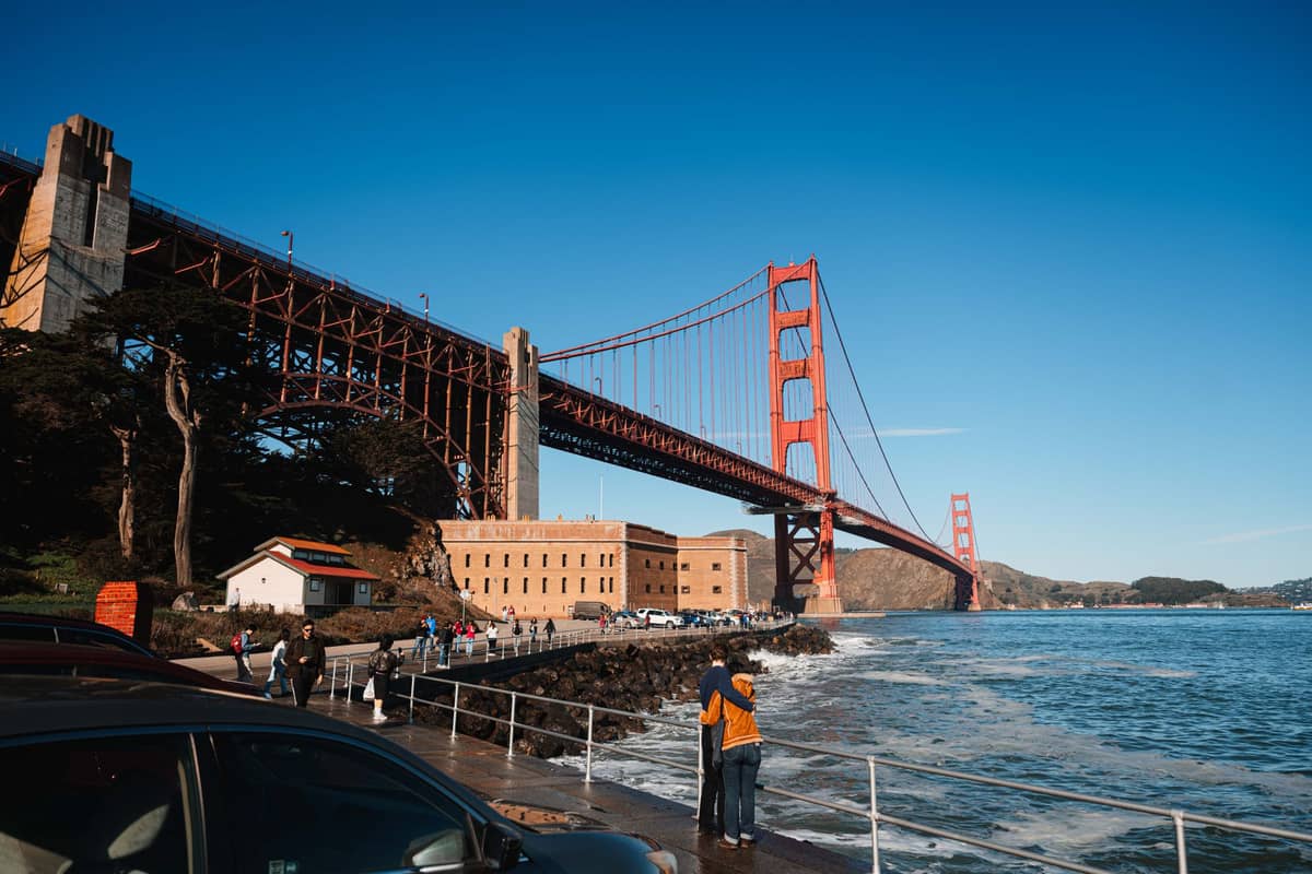 Vista del puente Golden Gate y Fort Point en San Francisco, con autos estacionados y turistas caminando por el paseo marítimo en un día soleado.