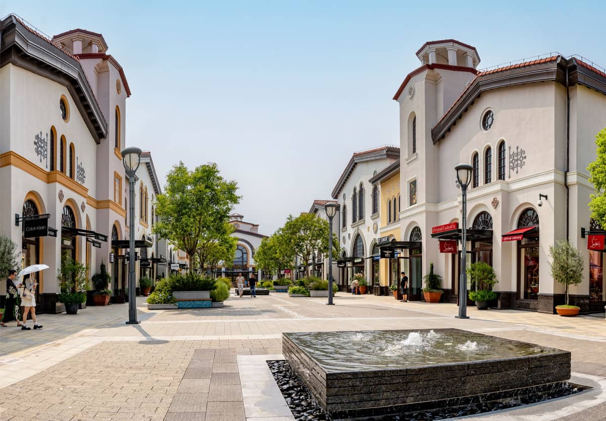 Area outlet di lusso con edifici in stile europeo, alberi, vetrine e una fontana centrale sotto cieli sereni.