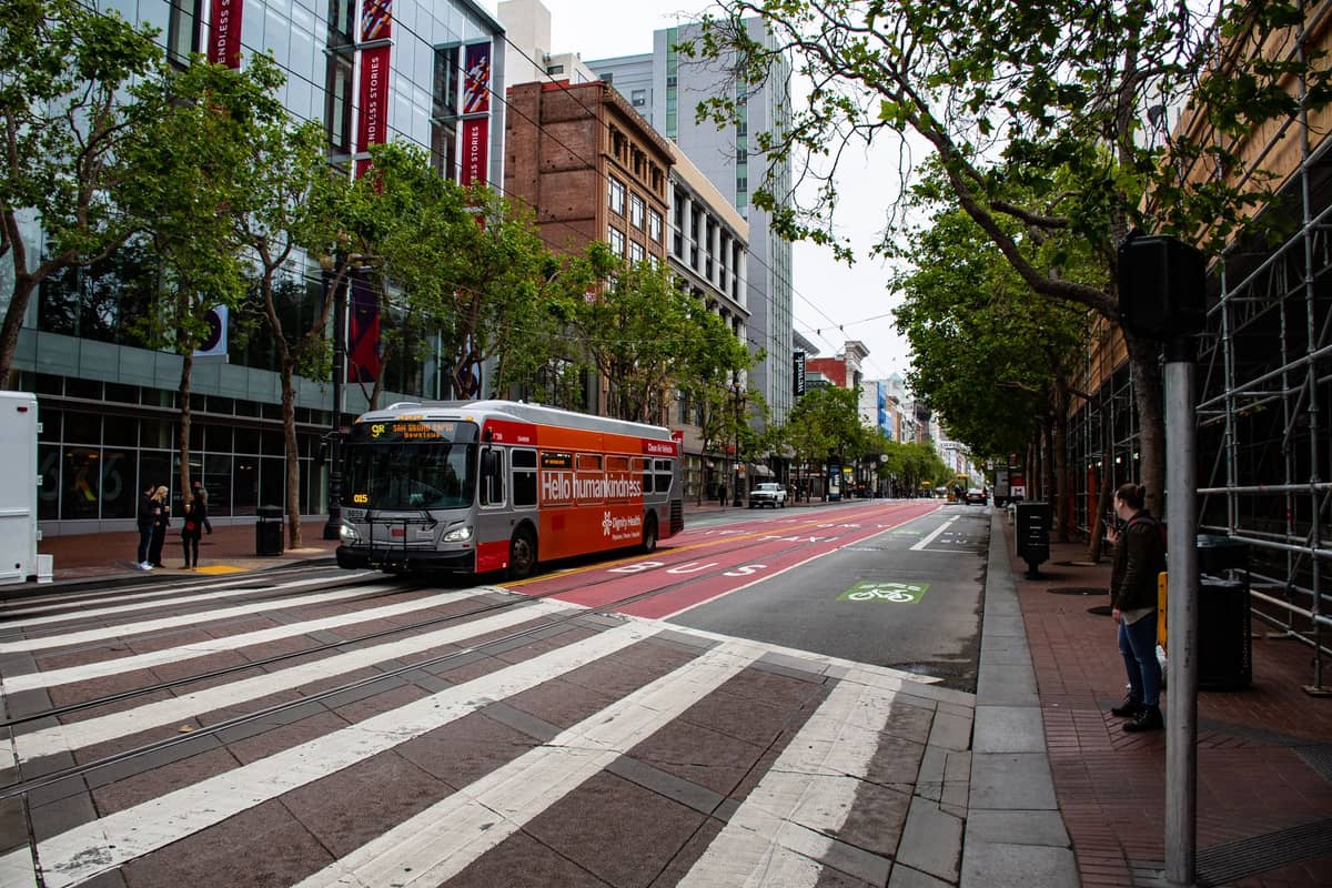 Eine Stadtstraße in San Francisco mit einem roten und silbernen Bus auf einer eigenen Busspur, Bäumen und hohen Gebäuden entlang der Straße.
