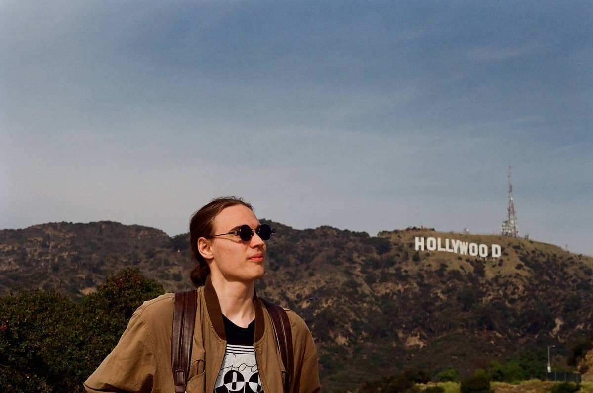 Un viajero con gafas de sol y mochila está de pie frente al icónico Hollywood Sign en Los Ángeles bajo un cielo despejado.