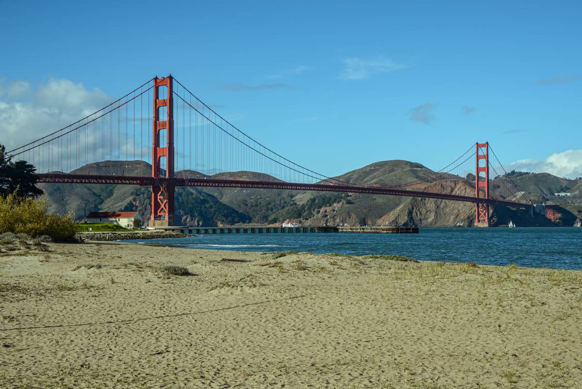 Puente Golden Gate visto desde una playa de arena del Presidio con cielo azul y agua en primer plano.