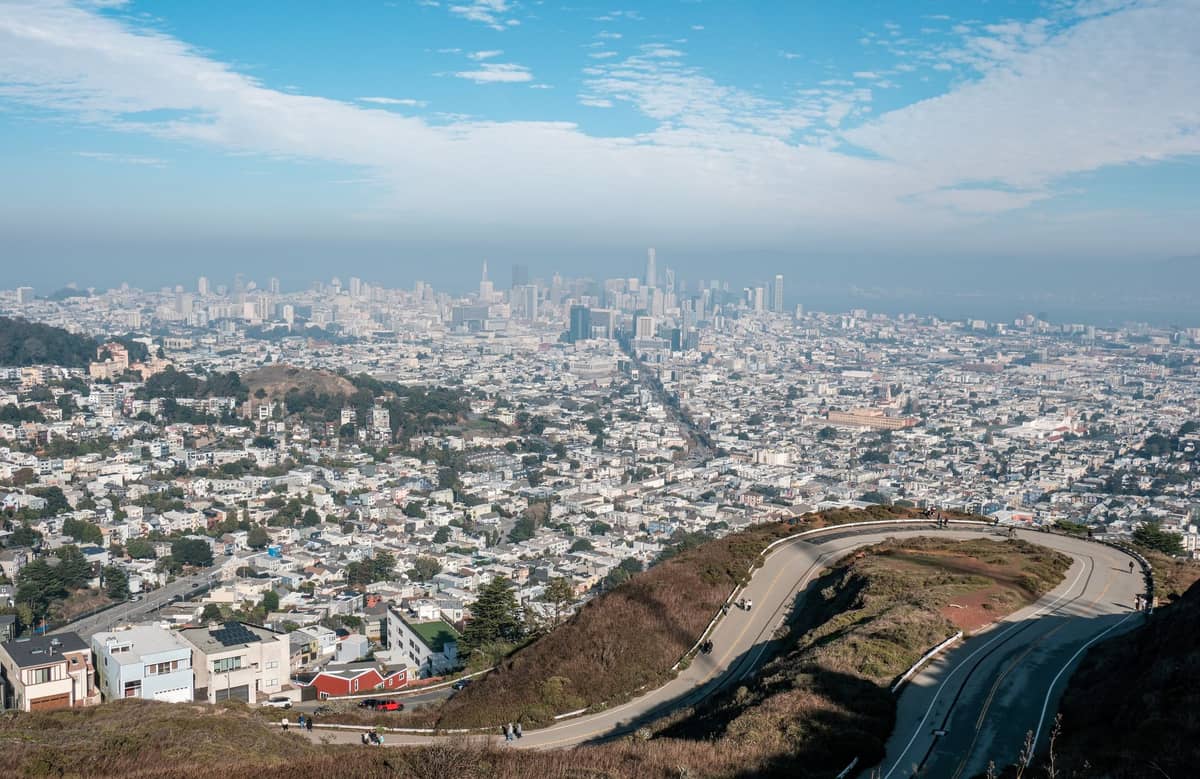 Vista panorámica diurna del horizonte de San Francisco desde Twin Peaks, con una carretera sinuosa en primer plano y la ciudad extendiéndose hasta el horizonte bajo un cielo azul.