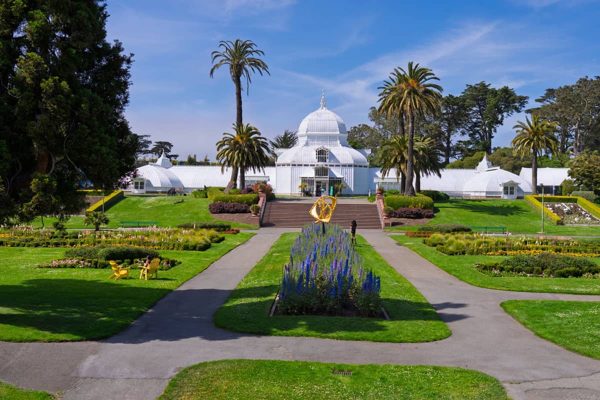Vista amplia del Conservatory of Flowers de San Francisco con jardines formales y palmeras bajo un cielo azul en Golden Gate Park.