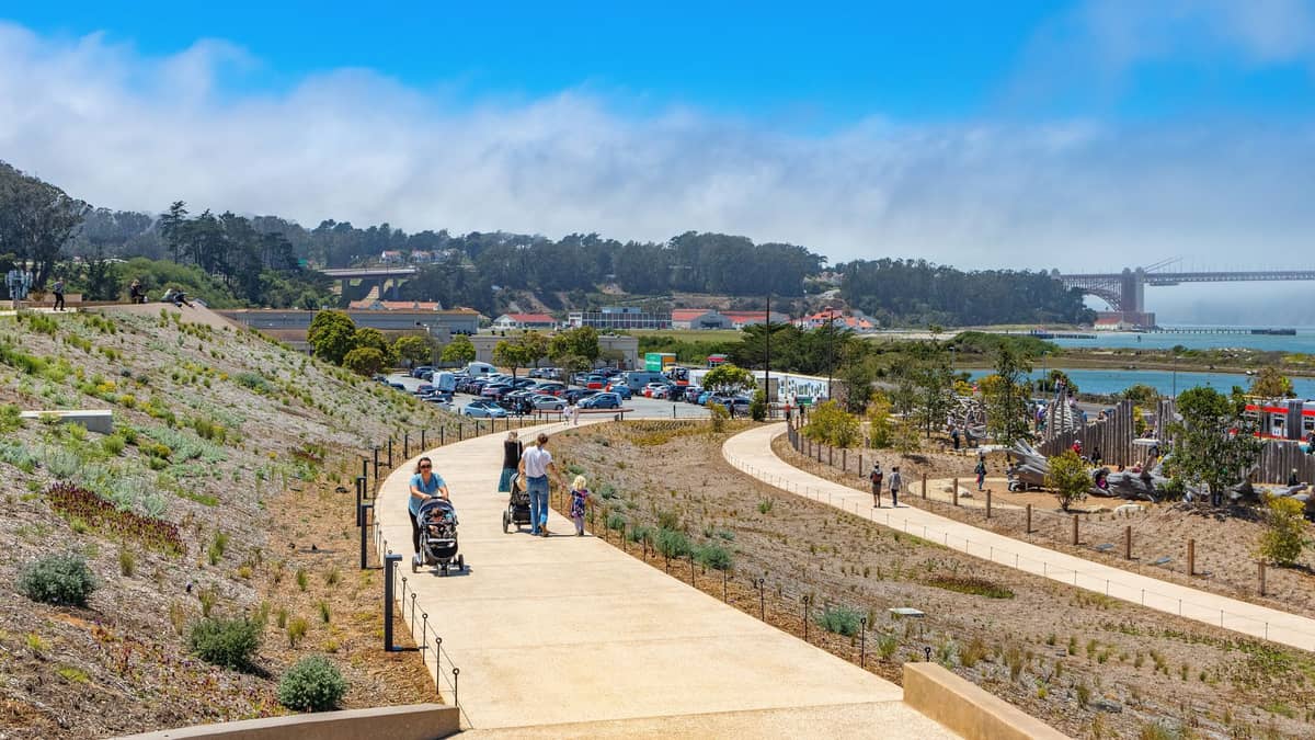 Familias con niños y carriolas pasean por un amplio sendero pavimentado en el Presidio de San Francisco, con el puente Golden Gate y la bahía visibles al fondo.