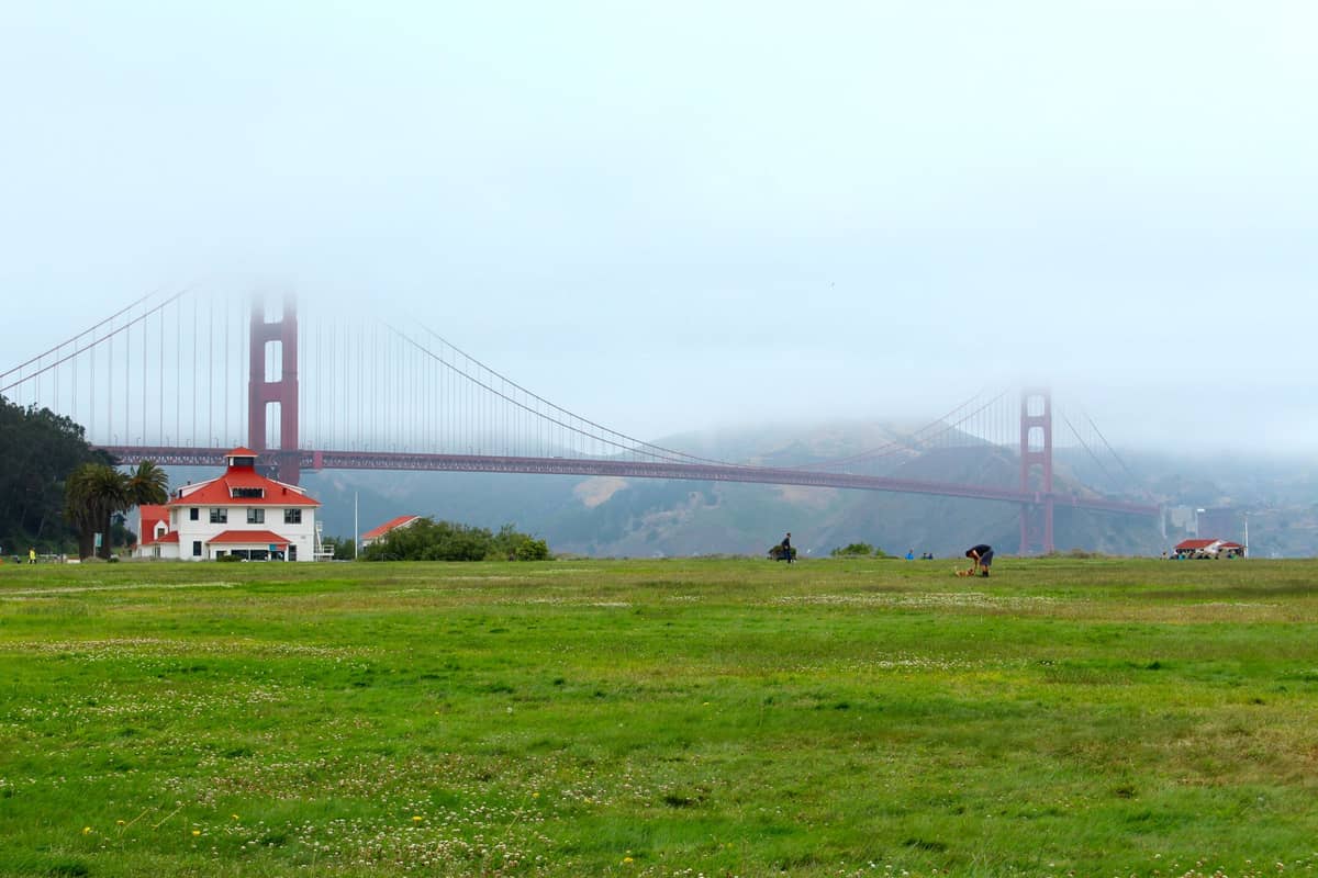 Blick auf die Golden Gate Bridge mit ihren Türmen im Nebel, gesehen über ein großes Grasfeld und ein weißes Gebäude im Vordergrund.