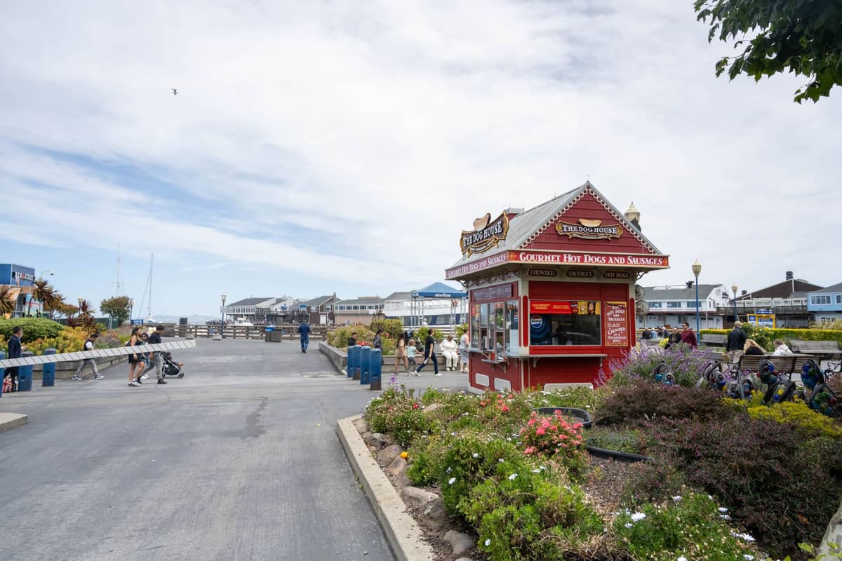 Pier-Bereich in San Francisco mit einem Hot-Dog- und Snackstand, Bänken, Spaziergängern und familienfreundlicher Umgebung unter blauem Himmel.