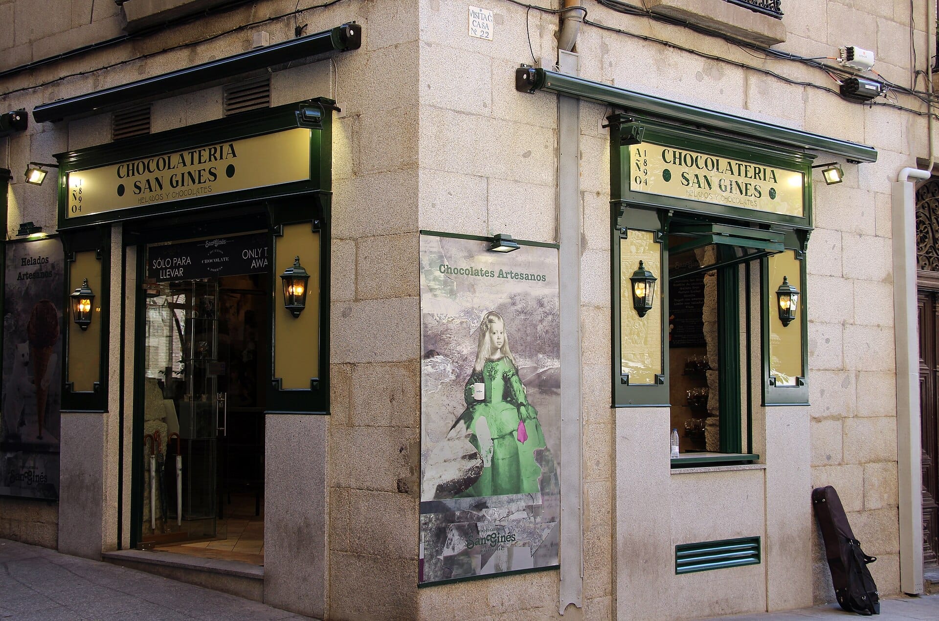 L'angolo esterno della Chocolatería San Ginés a Madrid, con la classica insegna verde, lanterne e un poster artistico vintage sulle pareti in pietra.
