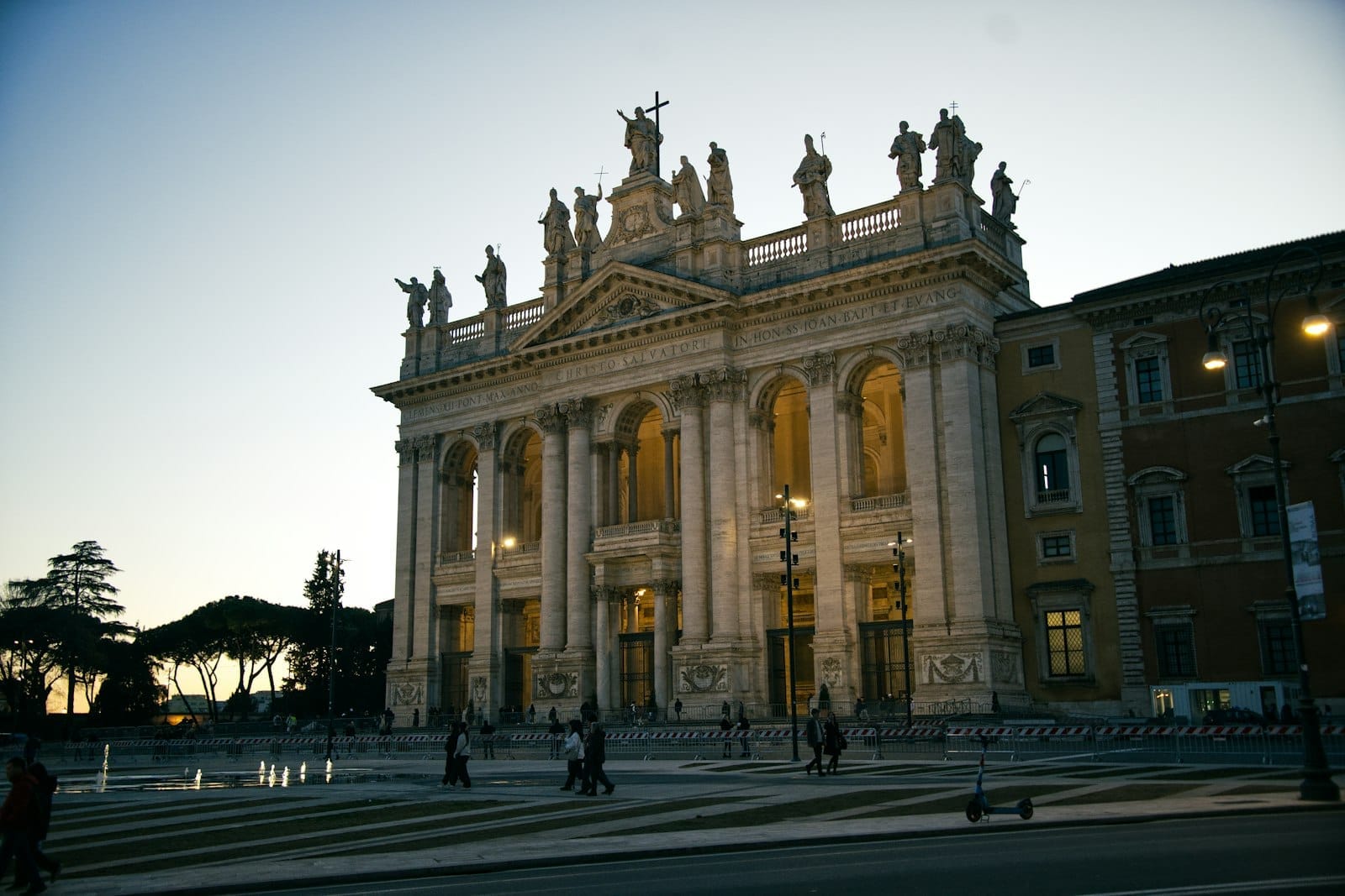 The illuminated facade of San Giovanni in Laterano basilica at dusk, with statues along the rooftop and people walking in the foreground.
