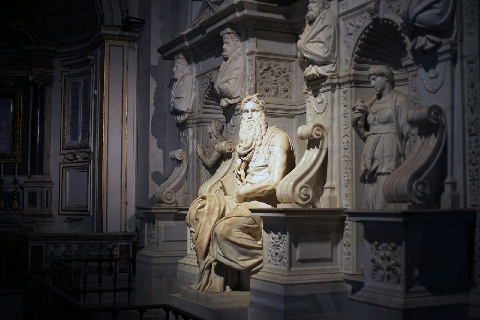 Michelangelo's Moses statue illuminated by soft light inside San Pietro in Vincoli, surrounded by intricate marble figures and architectural details.