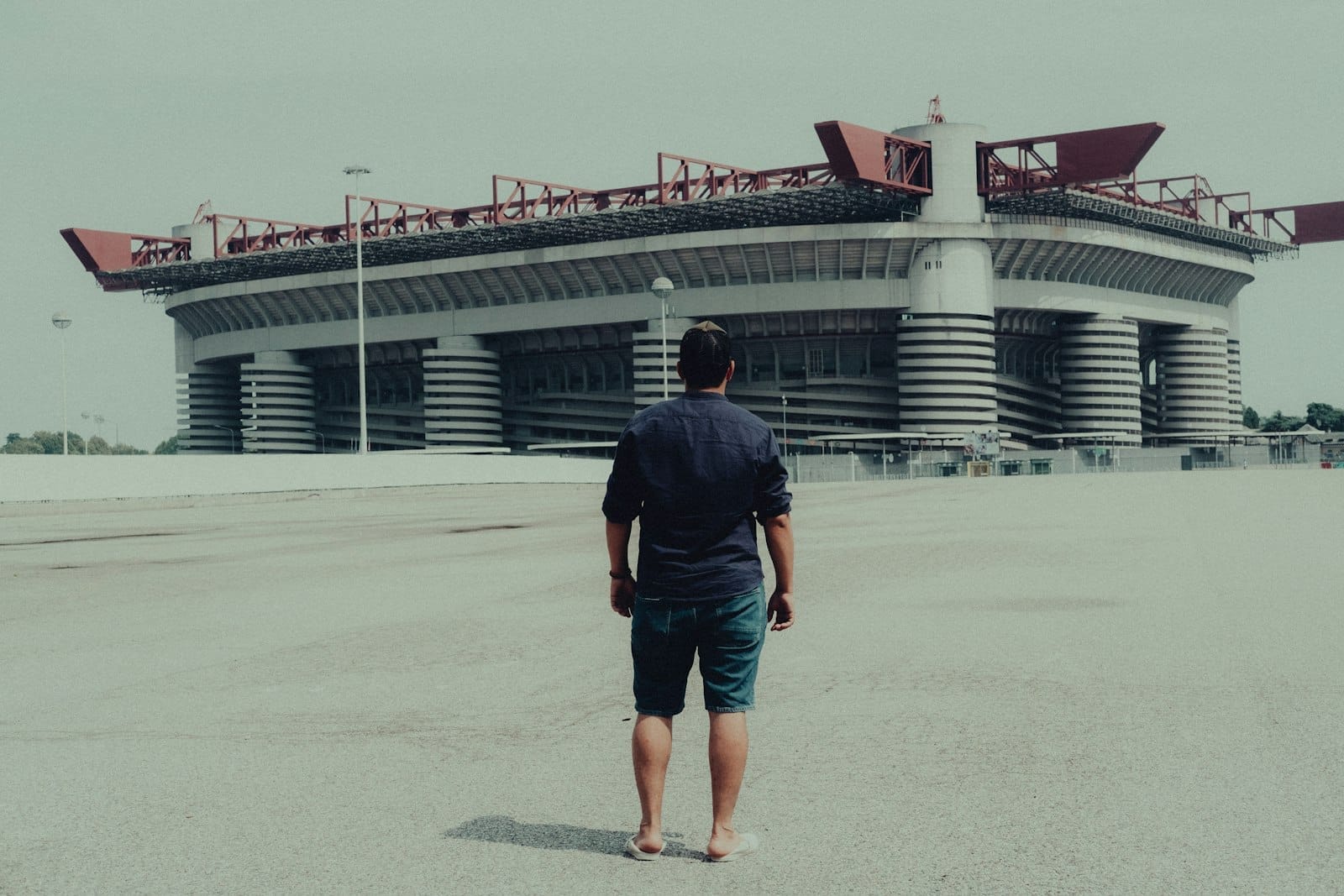 Una persona è in piedi su una piazza vuota davanti al maestoso Stadio San Siro, con il suo caratteristico tetto rosso e le colonne cilindriche.