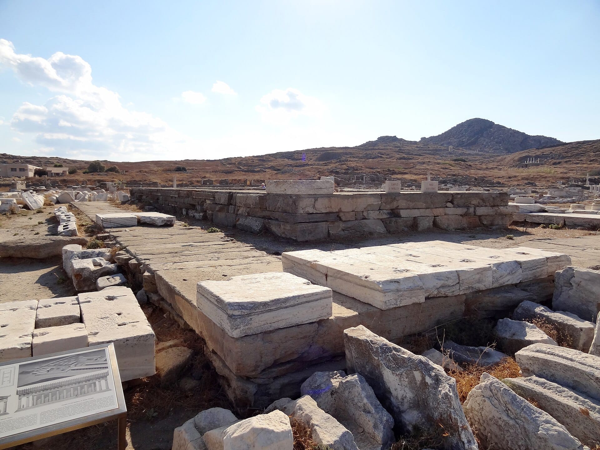 Ruinas de piedra antiguas del Santuario de Apolo en Delos, Grecia, rodeadas de un paisaje rocoso y un cielo azul despejado.