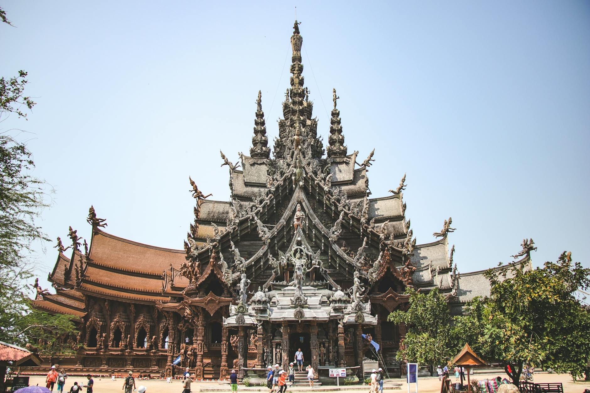 Vista ampia del tempio Santuario della Verità a Pattaya, con le sue imponenti guglie in legno e le decorazioni intagliate, sotto un cielo limpido e visitatori all'ingresso.