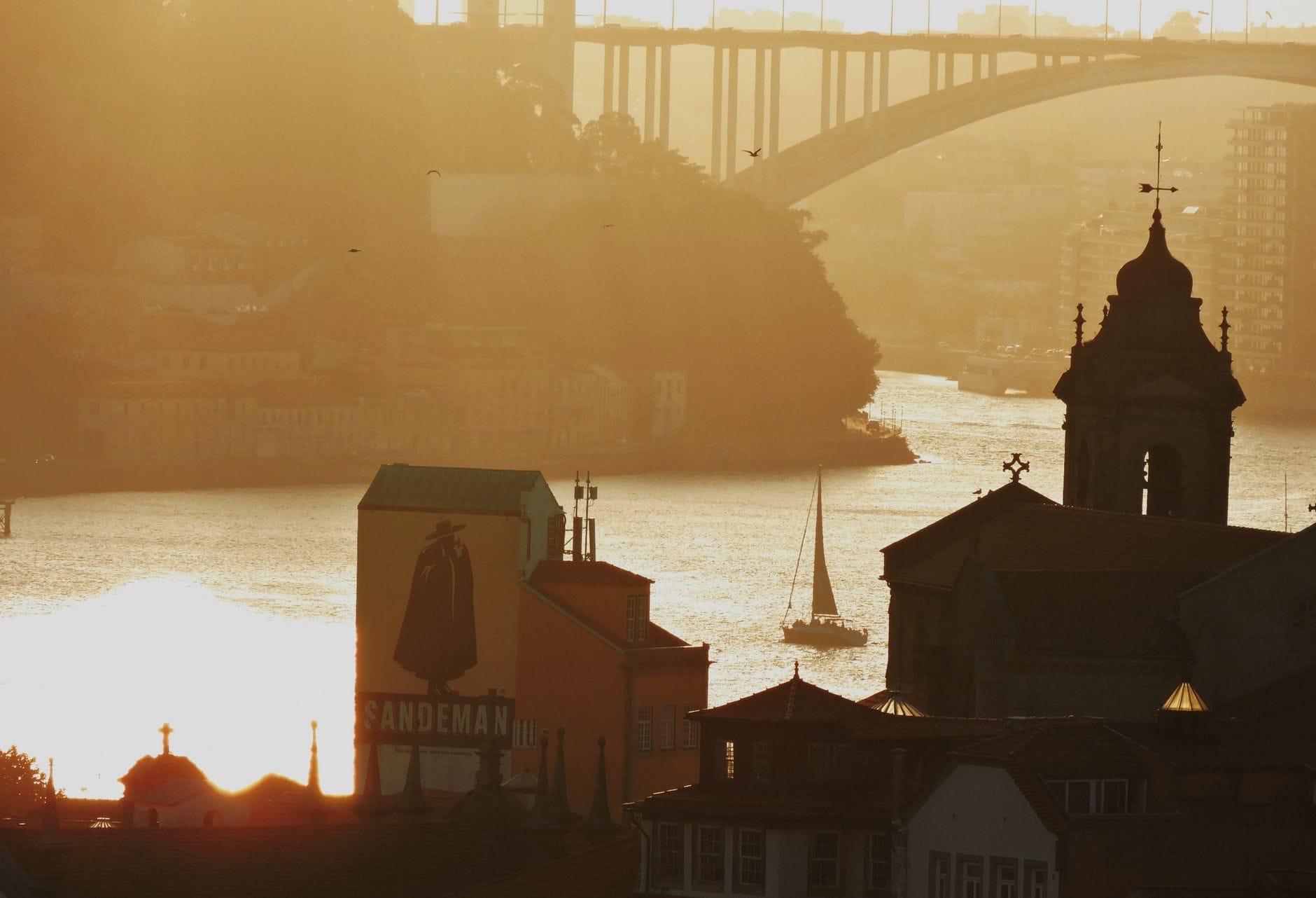 Vista al tramonto dorato sul fiume Douro con l'insegna delle cantine Sandeman, le barche vicine e l'iconico ponte di Porto.