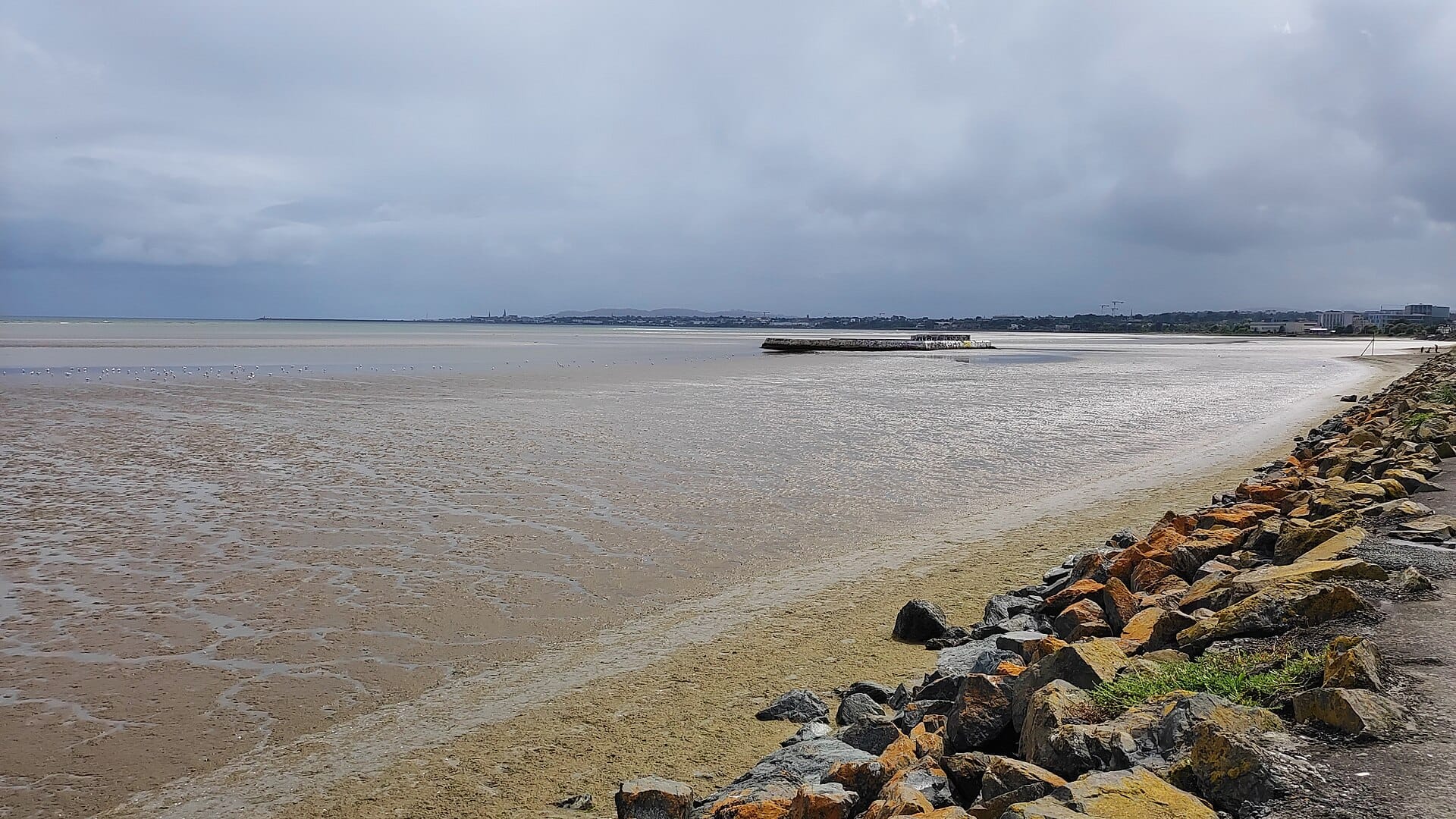 Sandymount Strand con la bassa marea, ampi distese sabbiose, riva rocciosa, un molo in lontananza e il cielo nuvoloso della Dublin Bay.