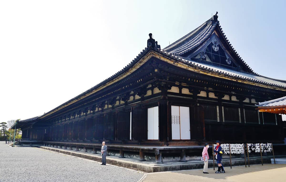 Wide-angle view of Sanjusangen-do Temple’s long wooden main hall with traditional tiled roof, against a clear sky, with a few visitors in front.