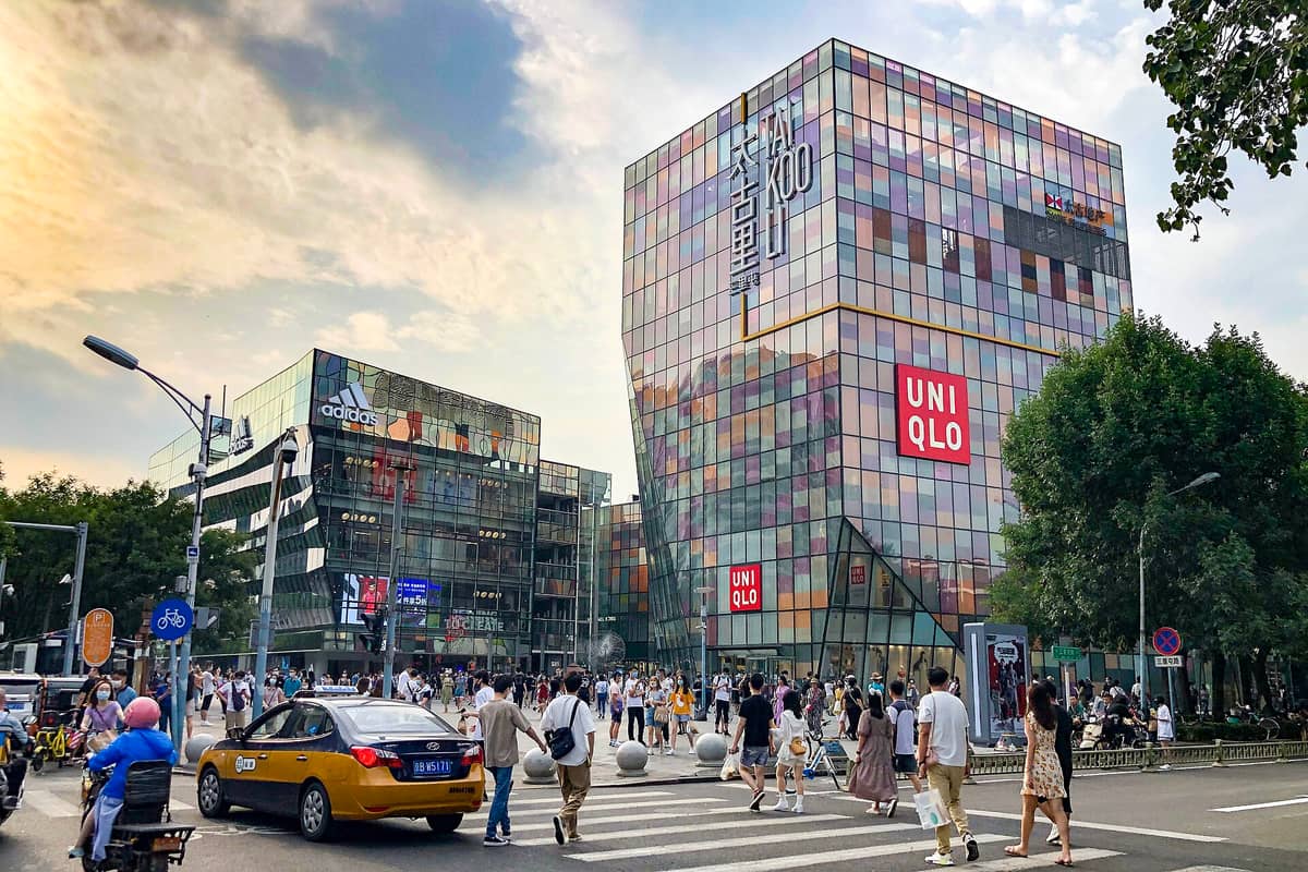 Belebte Straßenszene im Taikoo Li Sanlitun in Peking mit bunten Glasgebäuden, Uniqlo- und Adidas-Schildern, Menschenmassen, Bäumen und lebhaftem Stadtleben.