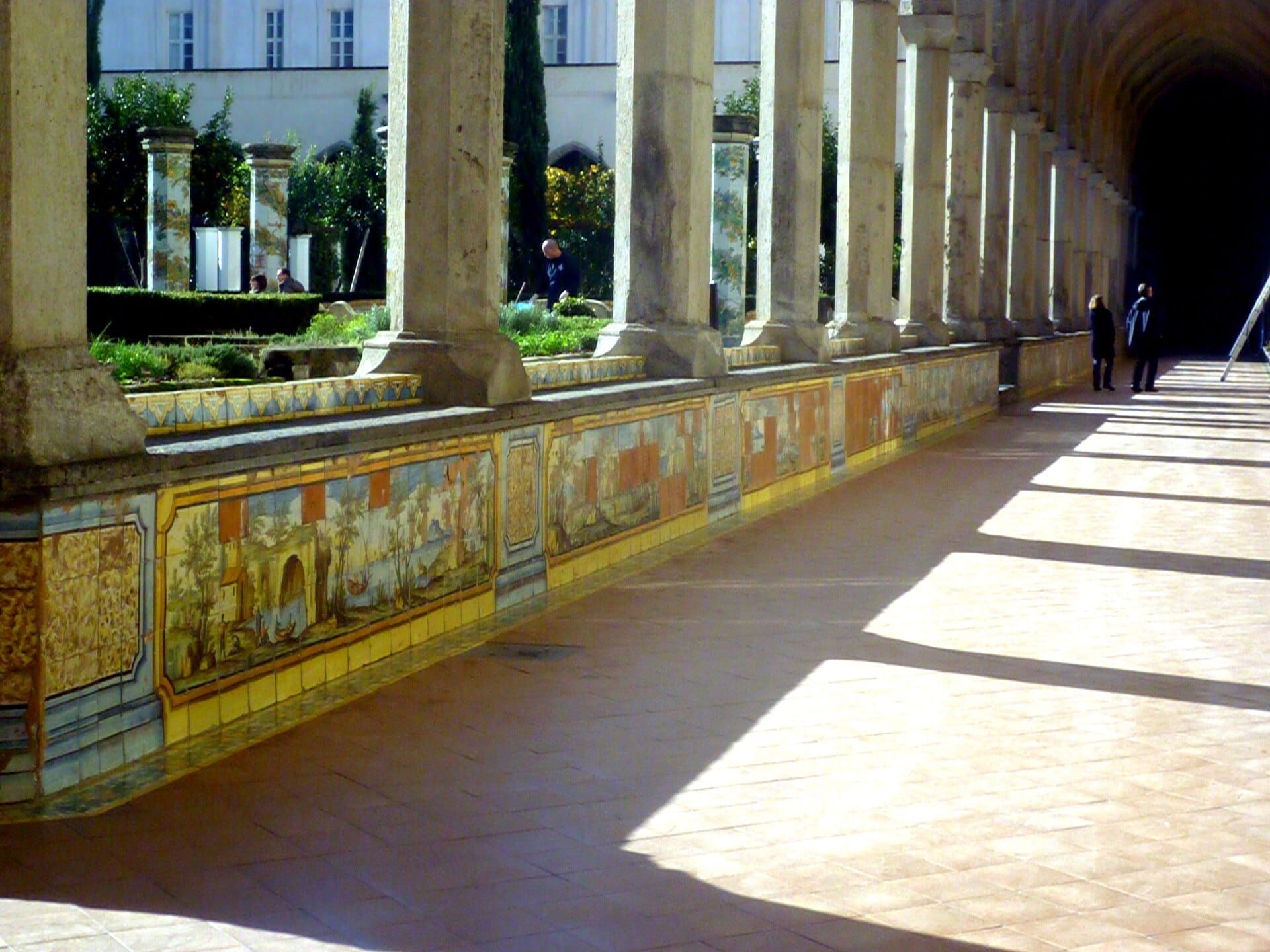 Pareti colorate con piastrelle di maiolica e colonne in pietra fiancheggiano il tranquillo corridoio del chiostro del Complesso Monumentale di Santa Chiara a Napoli sotto un sole brillante.