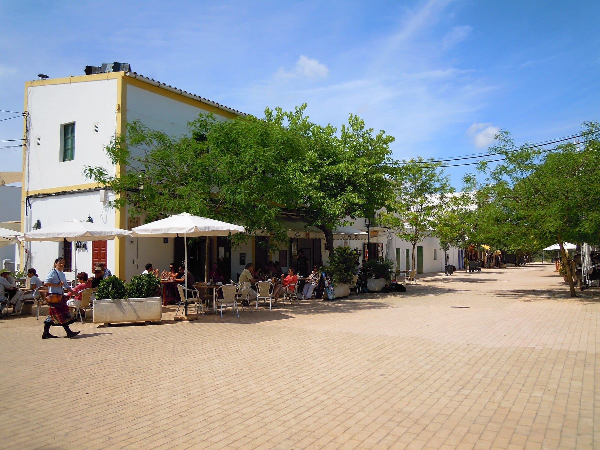Piazza del villaggio assolata a Santa Gertrudis de Fruitera, Ibiza, con edifici imbiancati, ombrelloni da caffè, alberi e persone che godono dei posti a sedere all'aperto.