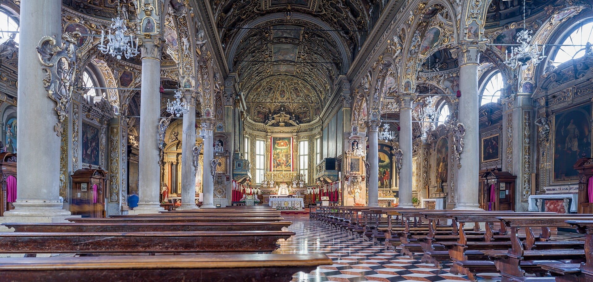 Vista interna di Santa Maria delle Grazie a Milano, con soffitti a volta, colonne decorative, lampadari e banchi in legno rivolti verso l'altare ornato.