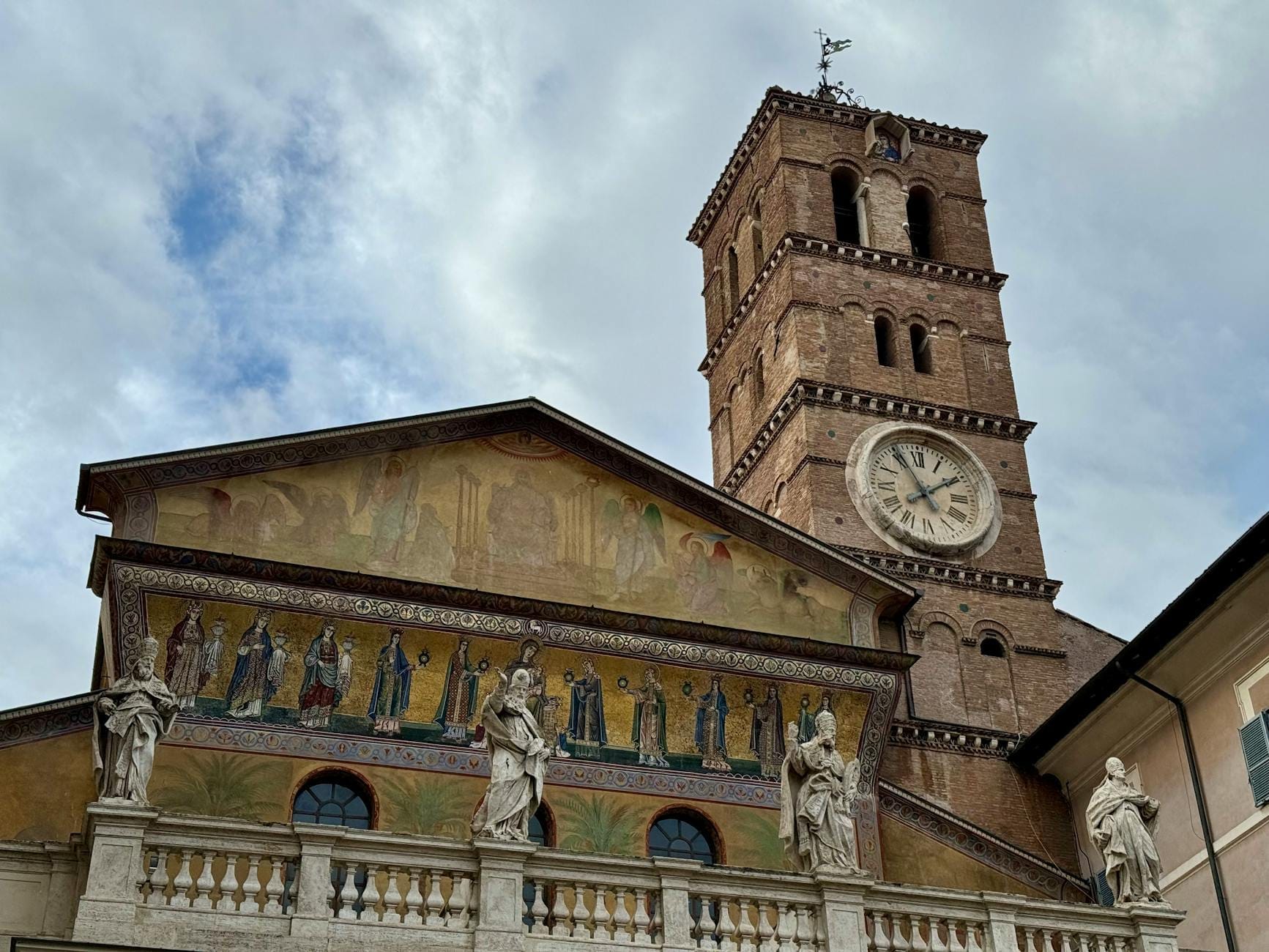 Facade of the Basilica di Santa Maria in Trastevere in Rome featuring golden mosaics, statues, and its distinctive bell tower under a partly cloudy sky.