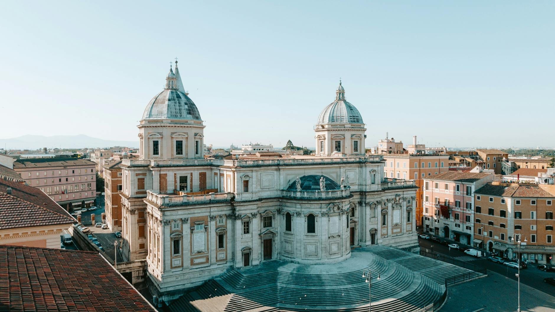 A wide view of the grand facade and twin domes of Santa Maria Maggiore basilica in Rome, with broad steps and cityscape in the background.