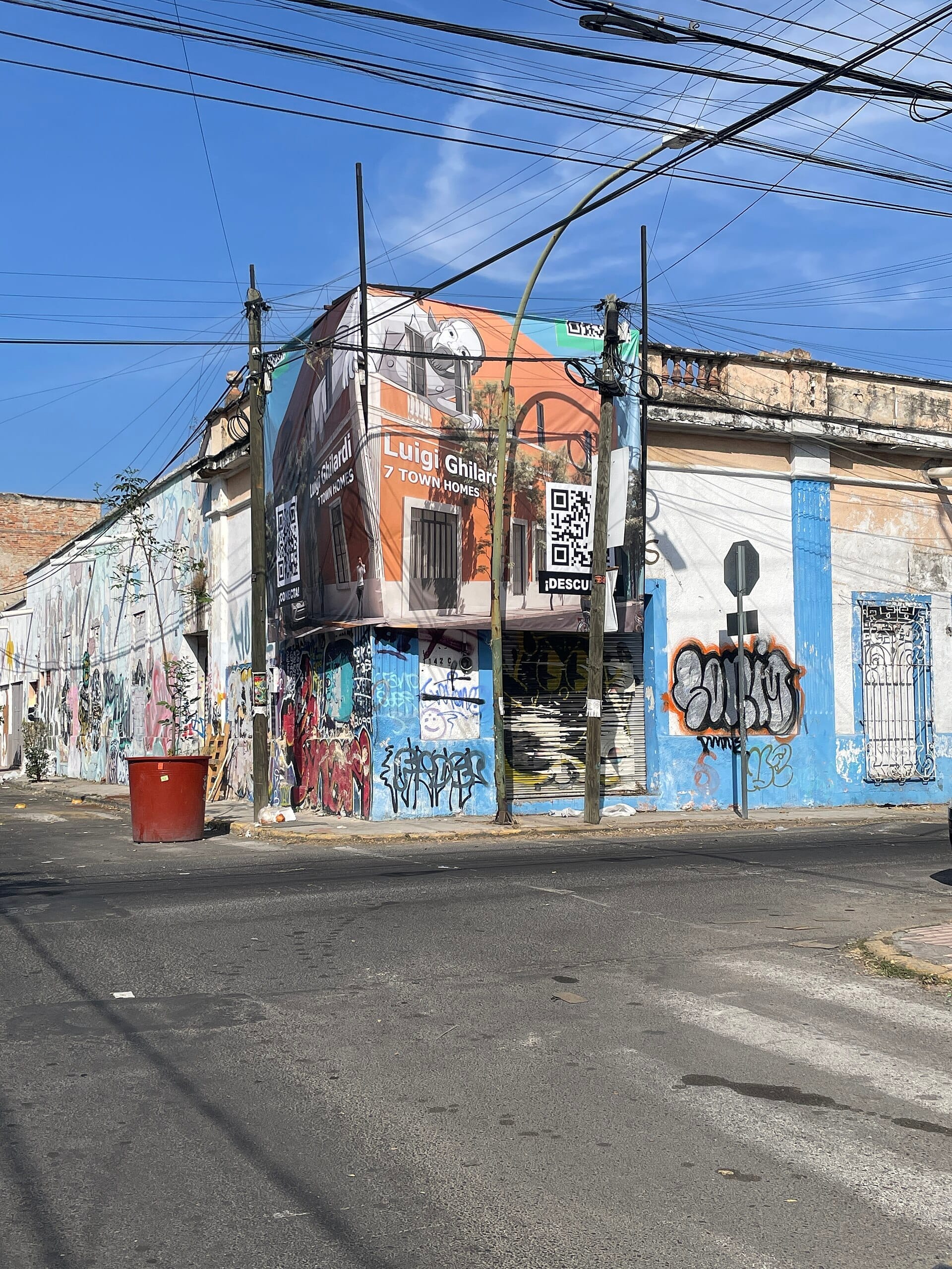 Angolo di strada a Santa Teresita, Guadalajara, con graffiti colorati, edifici antichi, un grande striscione pubblicitario e fili elettrici incrociati sotto un cielo azzurro limpido.