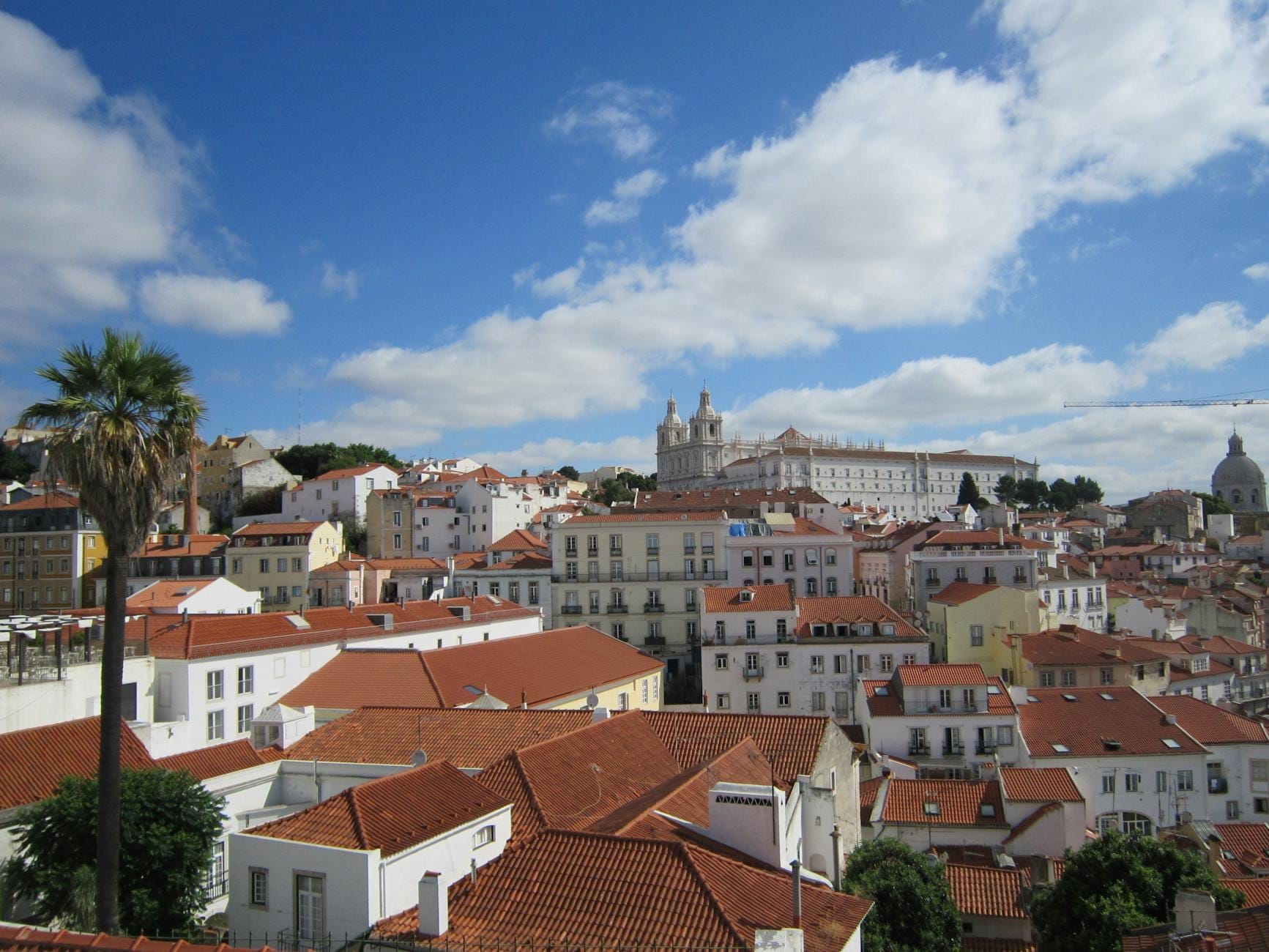 Vue sur le quartier historique d'Alfama à Lisbonne avec des toits rouges, un palmier et le monastère São Vicente de Fora sous un ciel bleu nuageux.