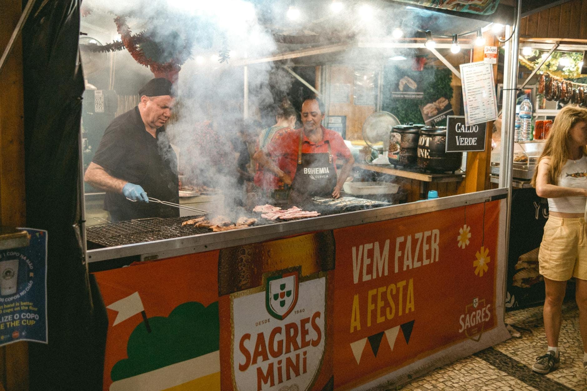 Deux personnes en train de griller des sardines dans un stand de restauration de rue animé avec de la fumée qui monte, des décorations de festival colorées et des azulejos portugais traditionnels.