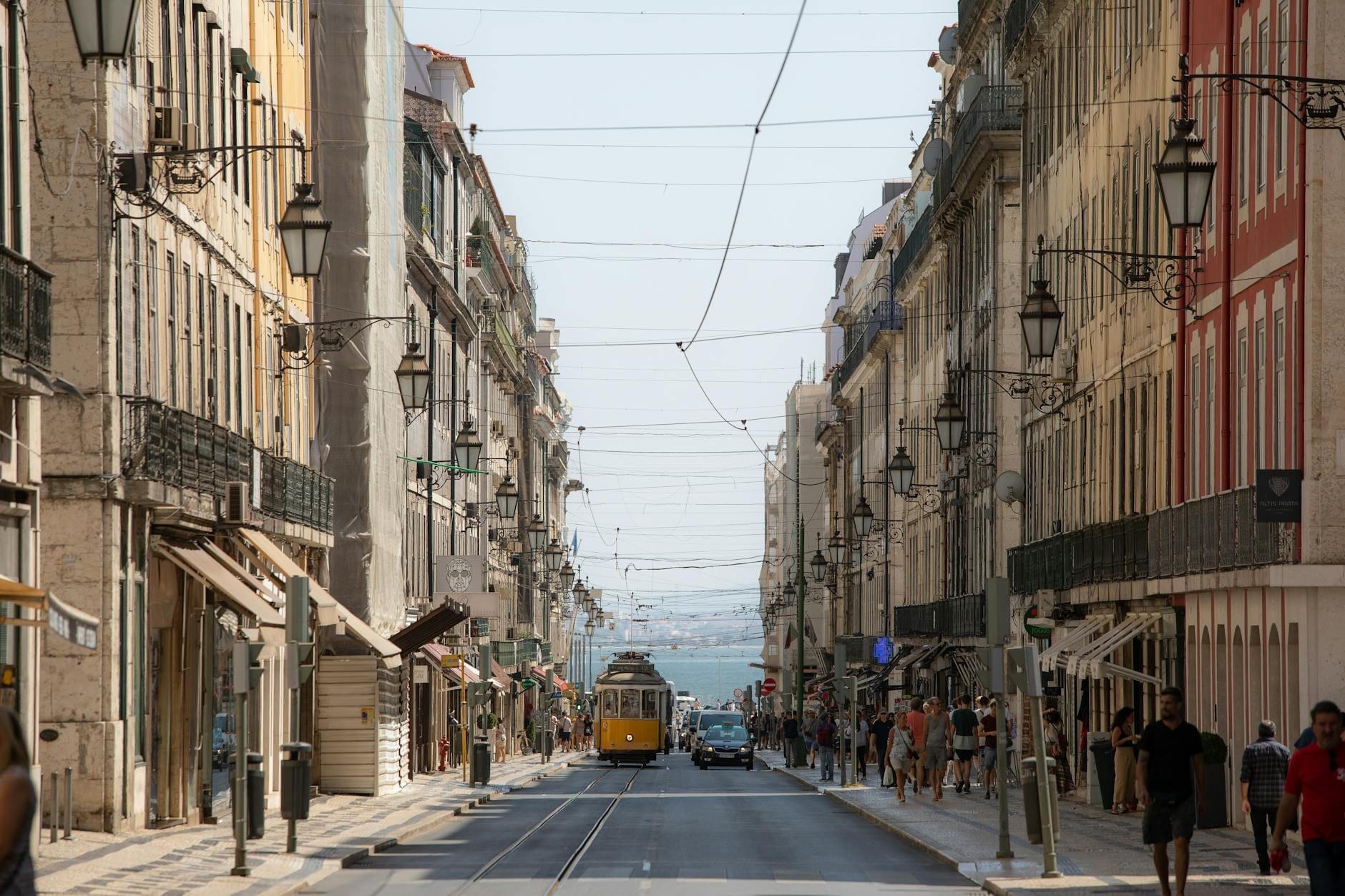 Large rue de Lisbonne avec un tramway classique, des bâtiments historiques et des guirlandes lumineuses festives suspendues par une journée ensoleillée.
