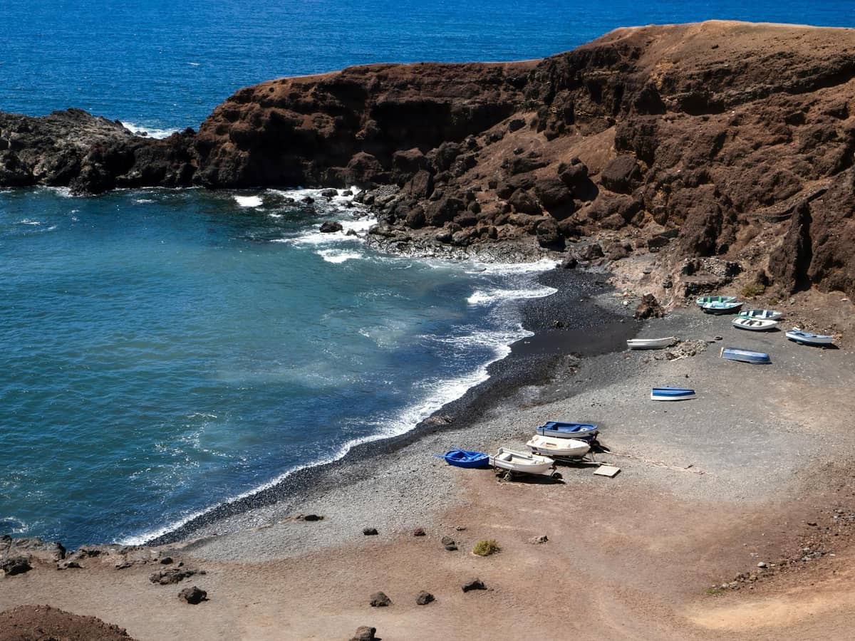 Geschwungener Schwarzsandstrand in Santorini mit kleinen Booten und vulkanischen Klippen rund um die Bucht bei hellem Sonnenlicht.