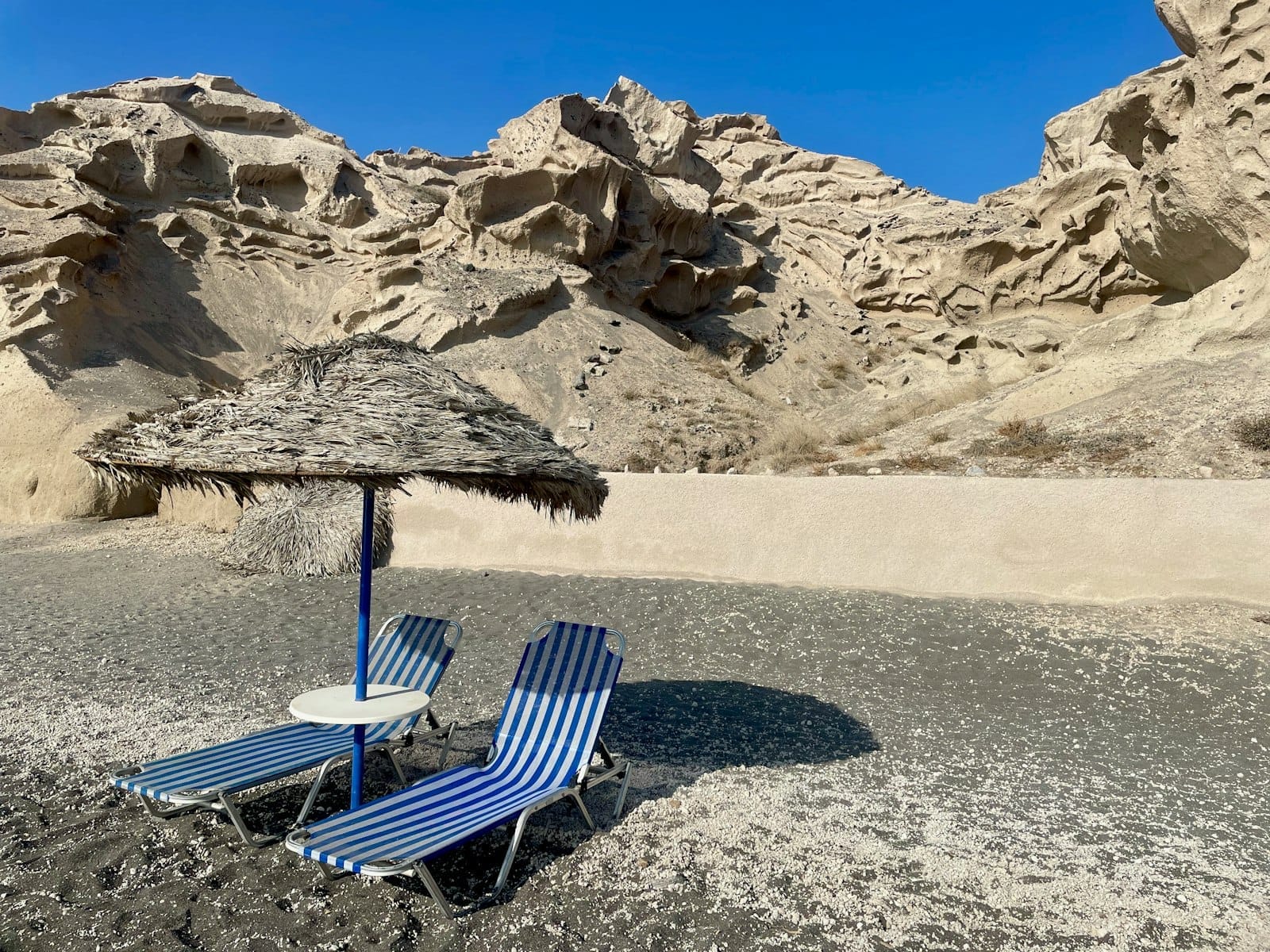 Duas espreguiçadeiras com listras azuis e um guarda-sol de palha em uma praia de areia negra em Santorini, com formações rochosas vulcânicas dramáticas ao fundo sob um céu azul.