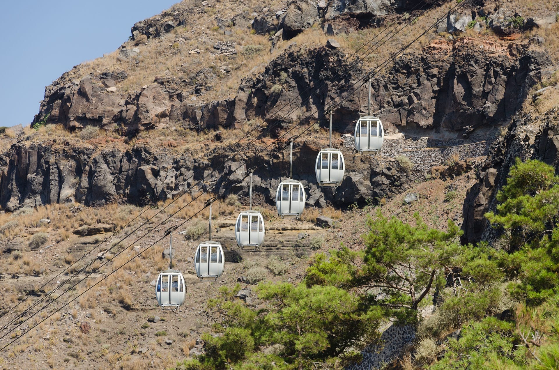 กระเช้าไฟฟ้า Santorini เคลื่อนที่ขึ้นไปตามหน้าผาหินที่ได้รับแสงแดด ซึ่งประดับด้วยหญ้าแห้งและพืชพรรณเบาบางใต้ท้องฟ้าสีฟ้าแจ่มใส
