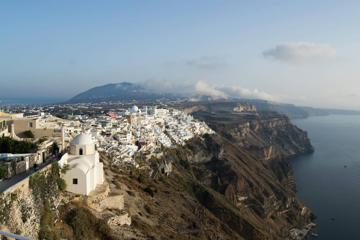 Panoramablick auf die weißen Gebäude von Firostefani, die auf den Klippen über der Caldera von Santorini thronen, mit dramatischer Küstenlinie und Meer darunter unter klarem Himmel.