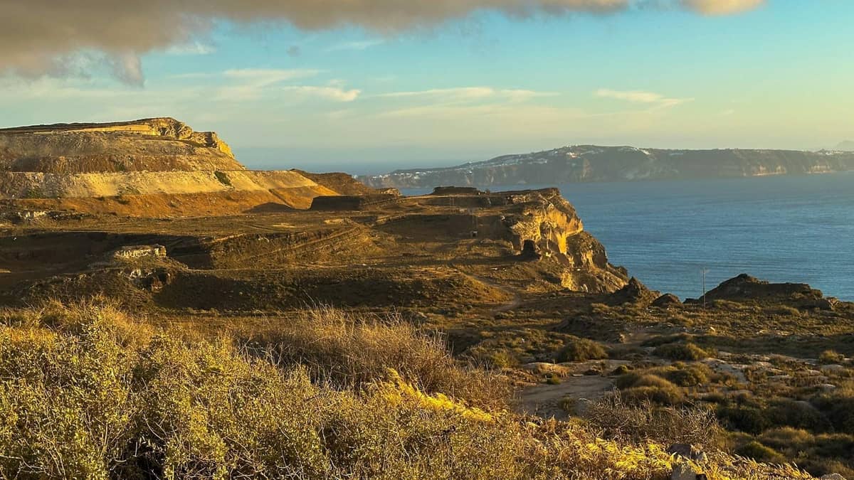 Paisaje costero de Santorini con acantilados escarpados, terreno amarillo-marrón, vegetación escasa y el Mar Egeo, evocando rutas hacia Akrotiri y Mesa Vouno.