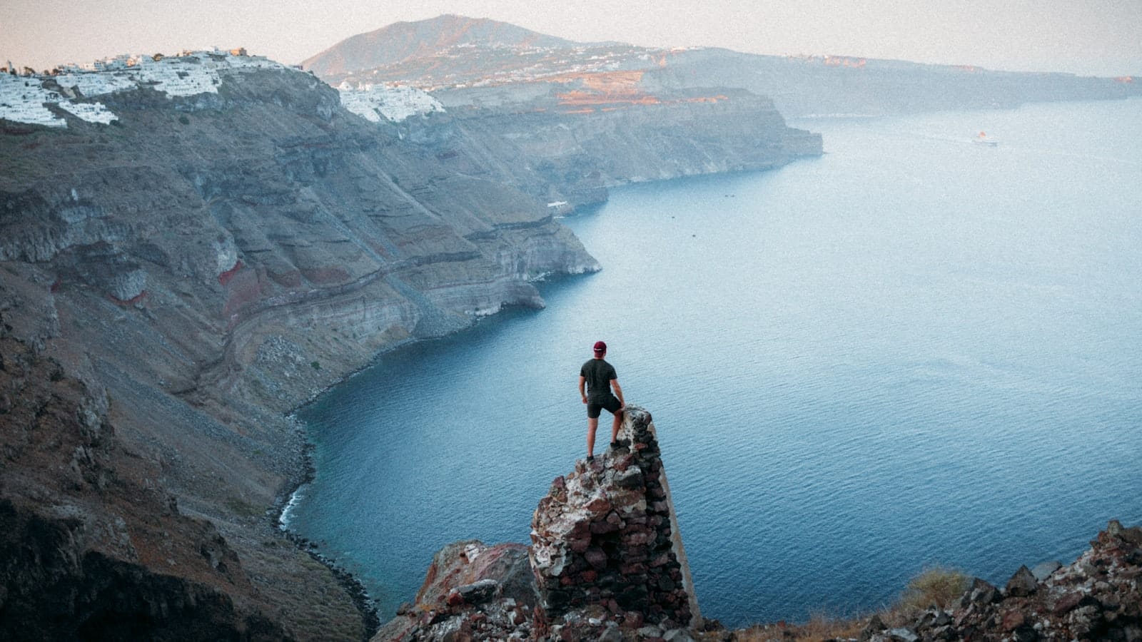 Um caminhante está em uma rocha com vista para as dramáticas falésias da caldeira de Santorini e o mar azul profundo ao pôr do sol, evocando aventura e uma paisagem deslumbrante.