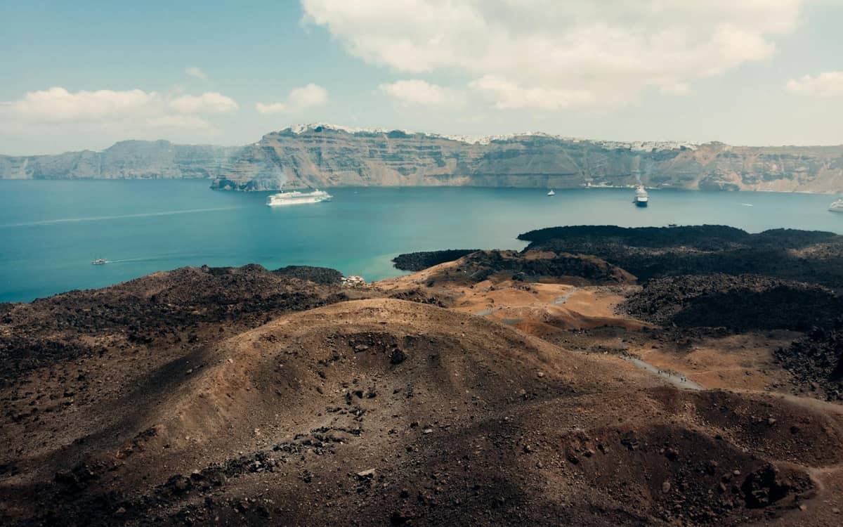 Terreno volcánico escarpado con rocas oscuras en primer plano, acantilados de la caldera y mar azul con cruceros en Santorini.