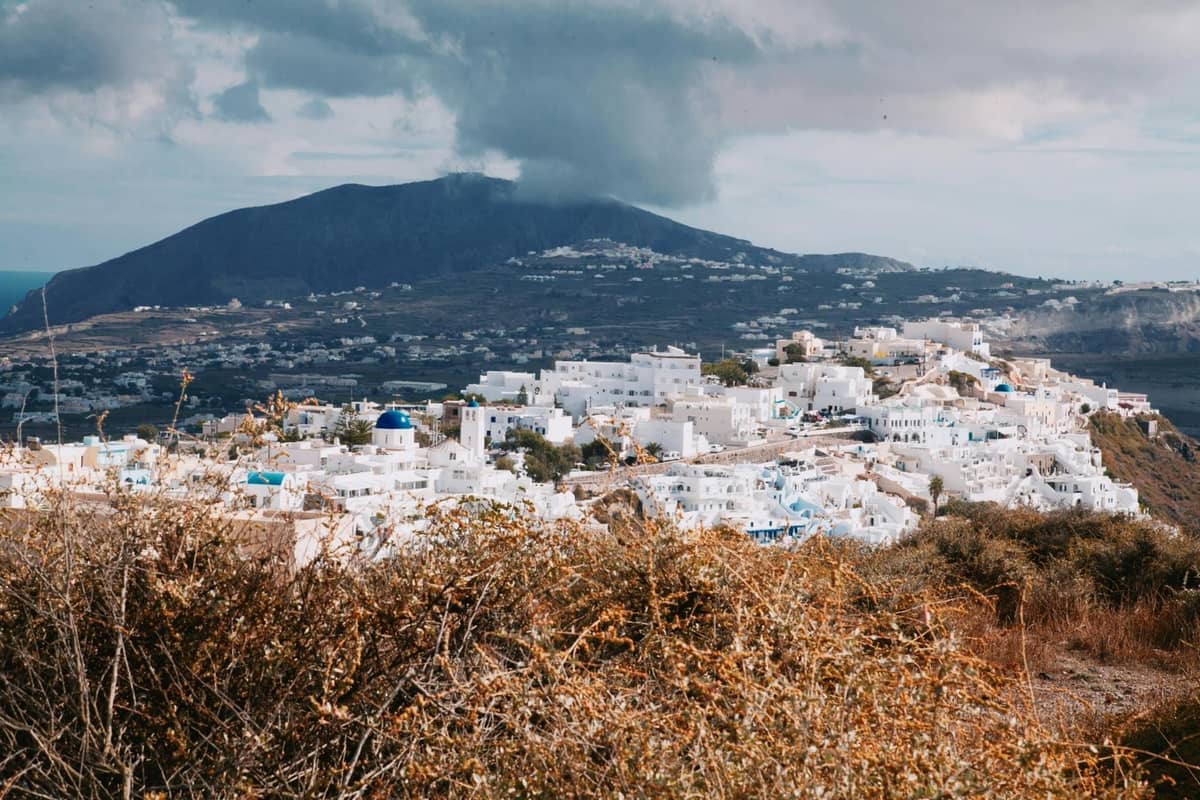 Pueblo en la cima de una colina en Santorini con edificios blancos y montañas de fondo, rodeado de paisaje rural y asentamientos tradicionales.
