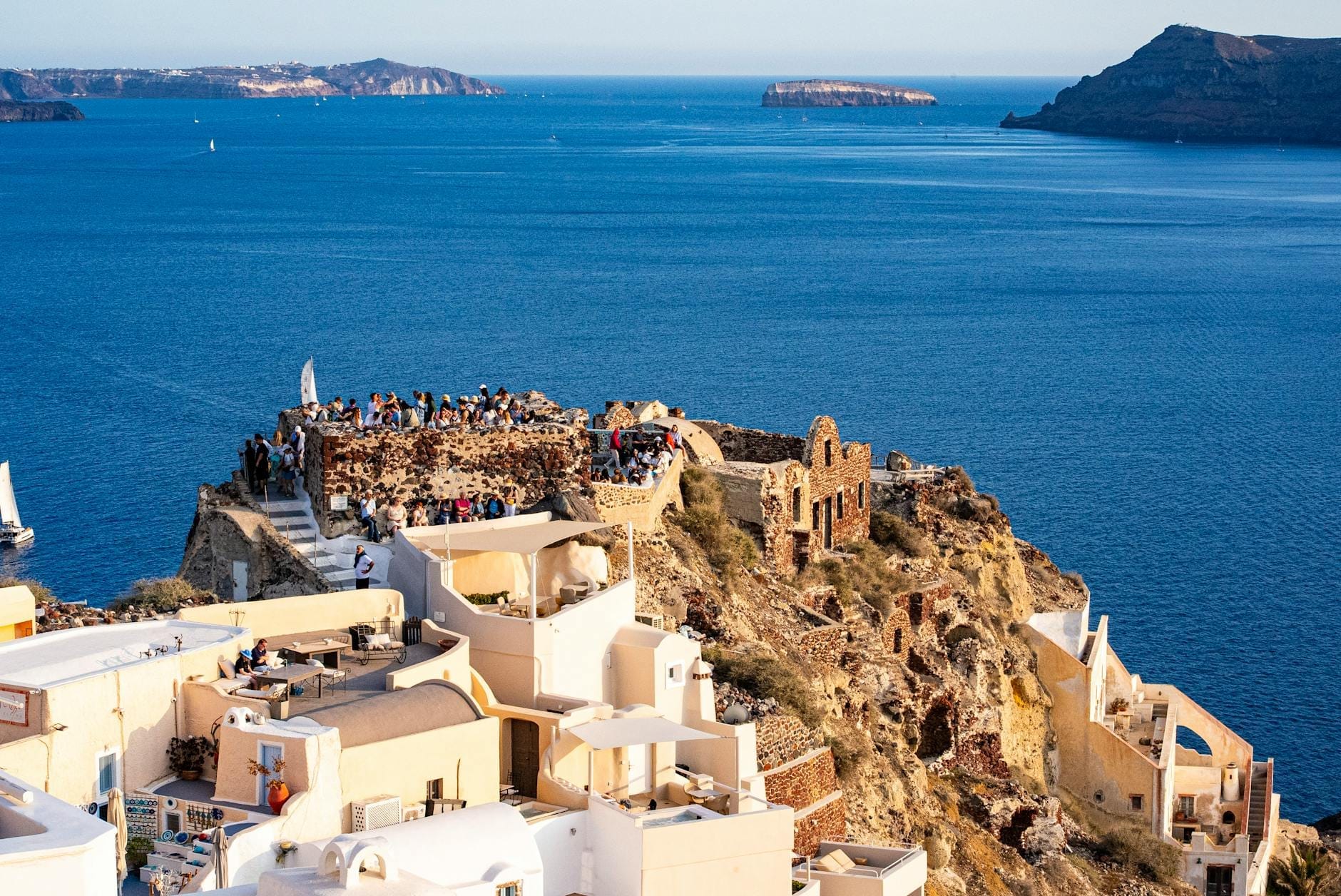 Vista das ruínas do castelo de Oia e multidões em um penhasco sobre o mar azul, com casas caiadas de branco em Santorini sob um céu limpo.