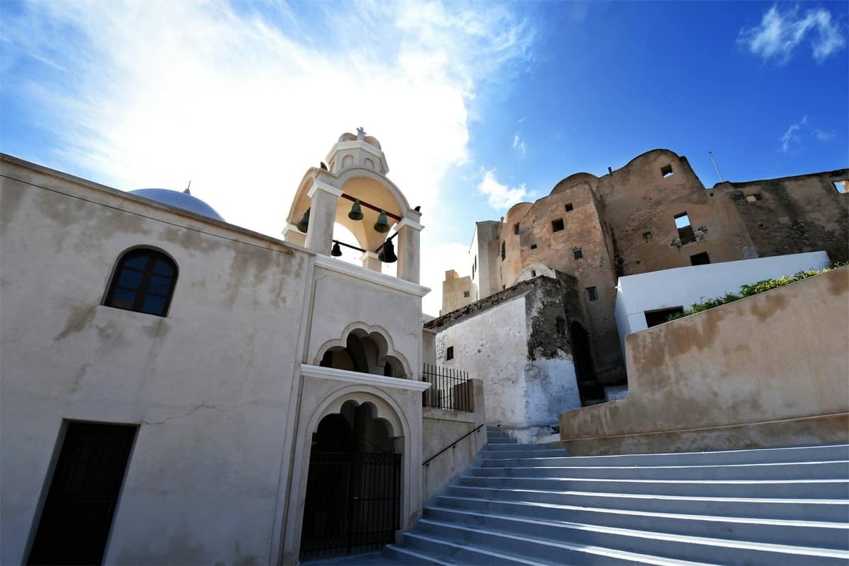 Traditionelles Gebäude mit gewölbtem Glockenturm und mittelalterlich wirkenden Steinruinen in Santorini unter einem strahlend blauen Himmel.