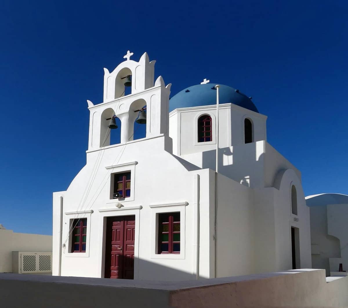 Weiß getünchte Kirche mit blauer Kuppel, drei Glockentürmen und roten Türen vor einem tiefblauen Himmel in Santorini.
