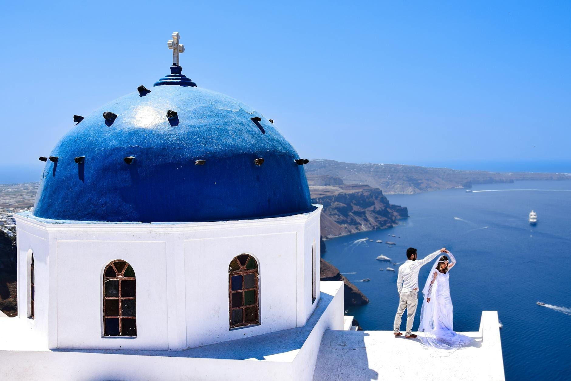 Noivos se abraçam em um terraço ao lado da icônica igreja de cúpula azul de Santorini, com as falésias da caldeira e o mar azul profundo sob um céu limpo.