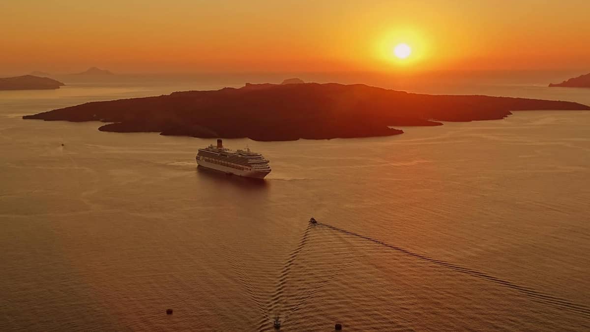 Großes Kreuzfahrtschiff in der Caldera von Santorini bei Sonnenuntergang mit der vulkanischen Insel als Silhouette vor einem orangefarbenen Himmel.