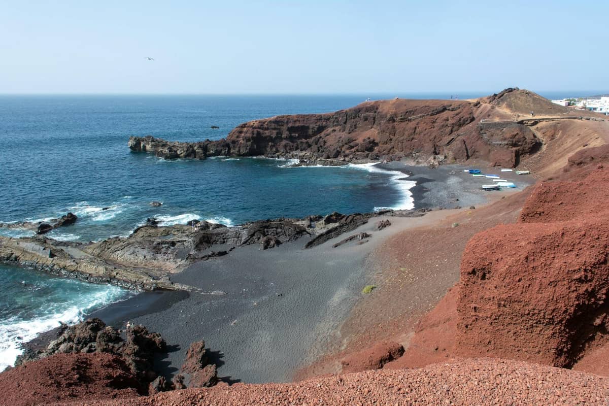 Küste von Santorini mit rostrot gefärbten Klippen und dunklem Vulkansandstrand, der auf das blaue Meer unter klarem Himmel trifft.