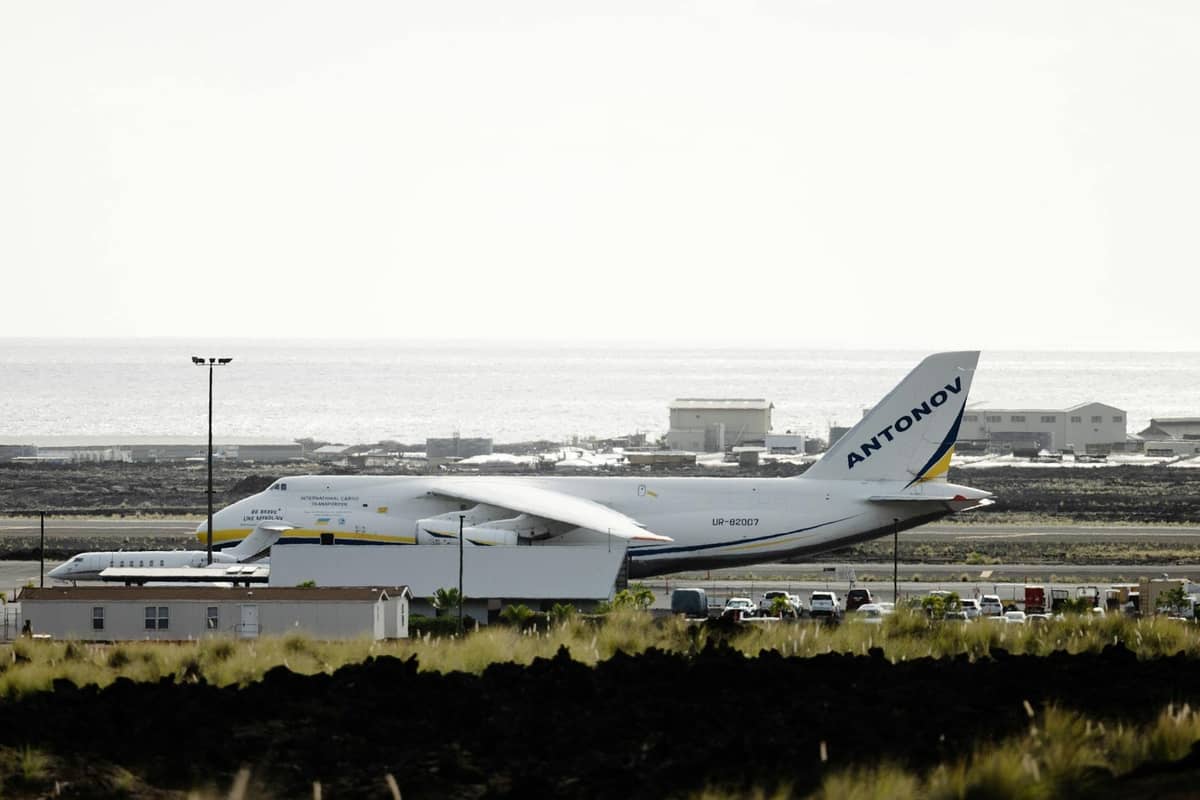 Un grande aereo Antonov parcheggiato in un piccolo aeroporto costiero con il mare e bassi edifici sullo sfondo.