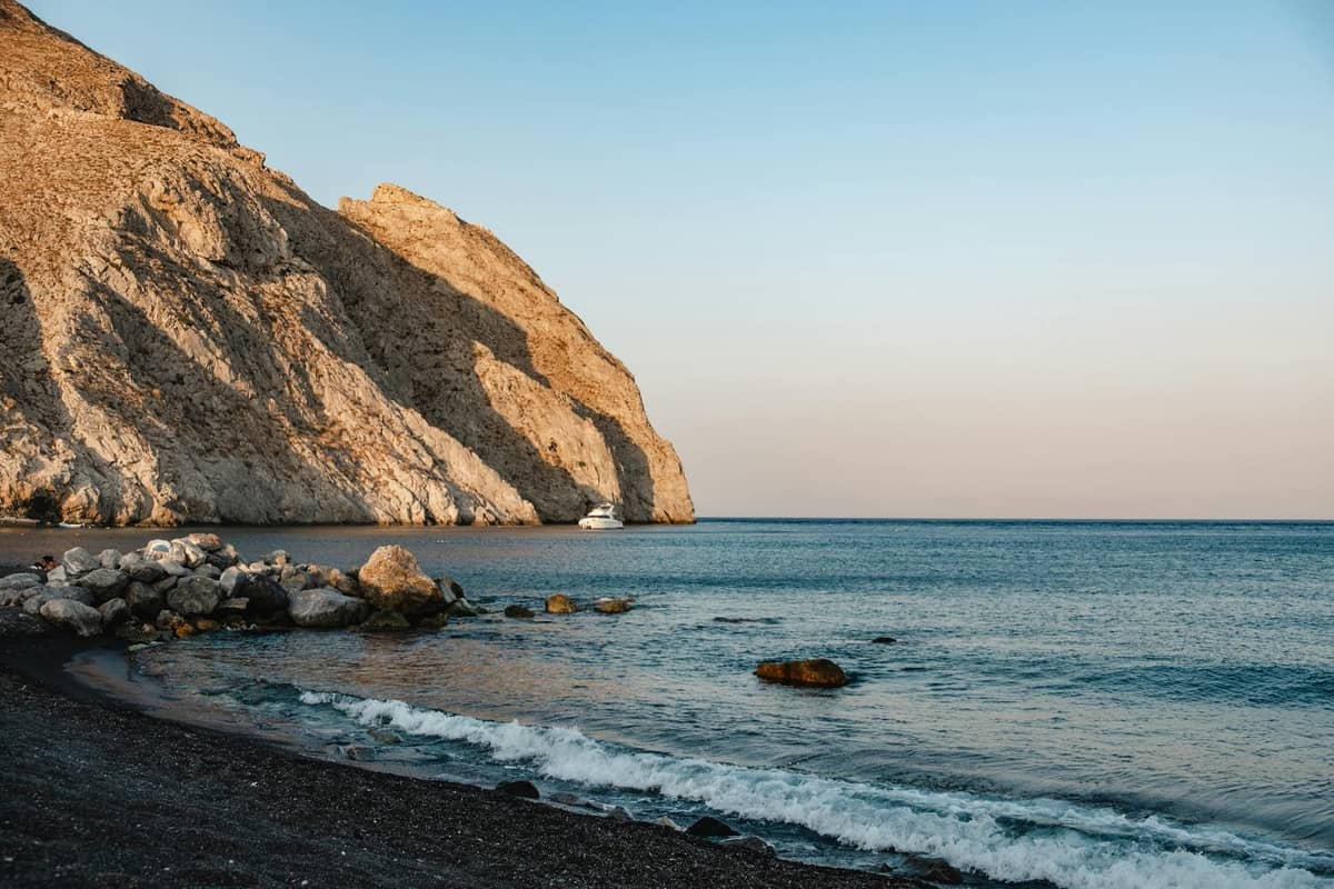 Una tranquilla spiaggia di Santorini con sabbia nera, onde gentili e spettacolari scogliere rocciose sotto un cielo sereno al tramonto.