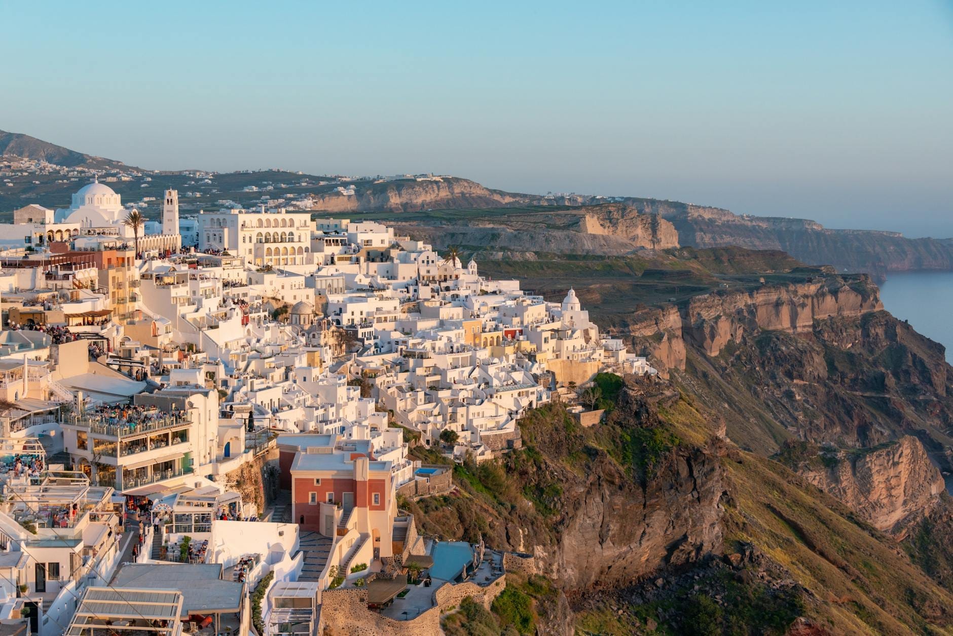 Vista panorâmica dos icônicos edifícios brancos de Santorini sobre as falésias dramáticas ao pôr do sol, com a caldera ao fundo banhada pela luz dourada característica de setembro.